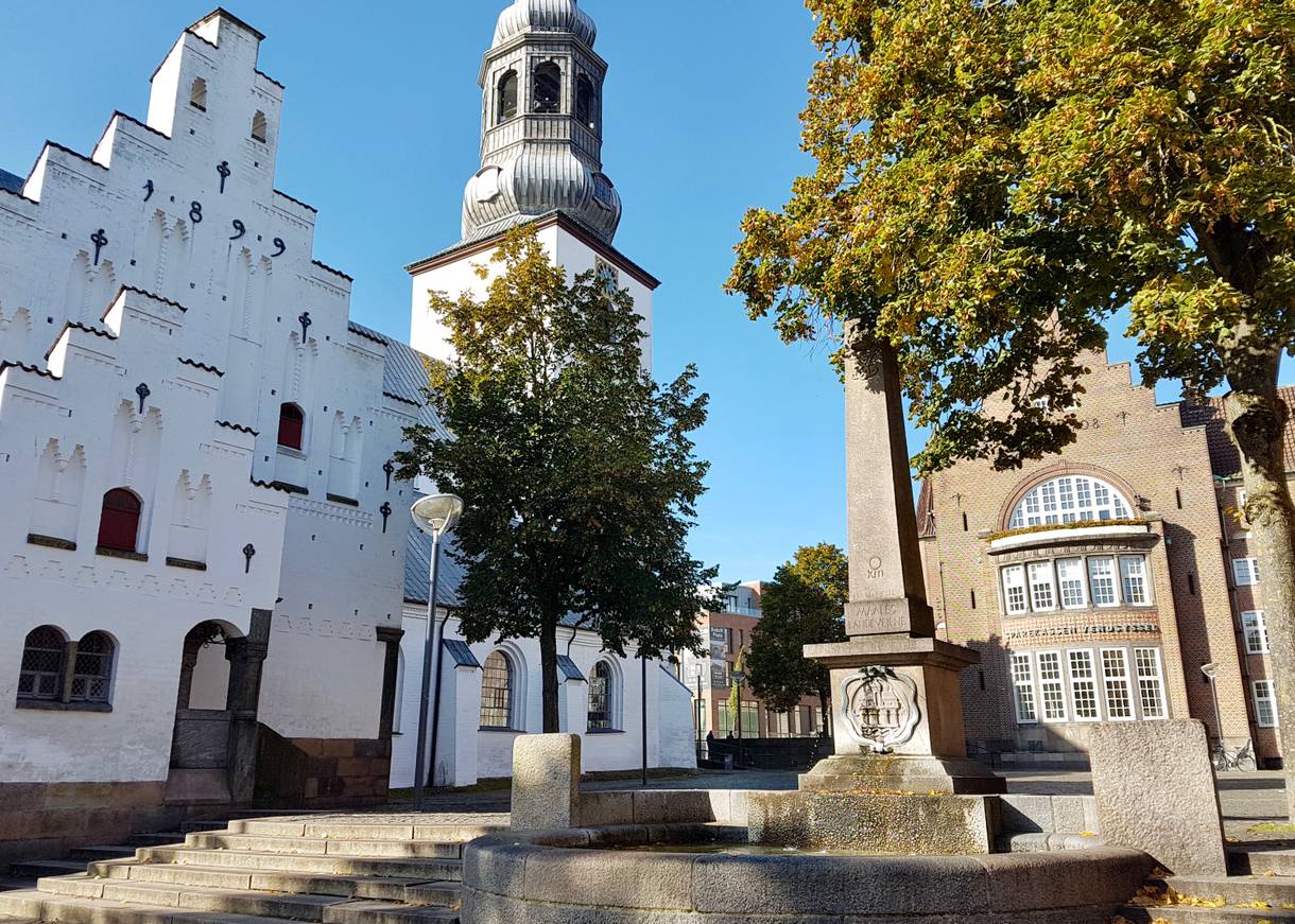 Obelist auf einem Sockel, dahinter die weiße Kirche