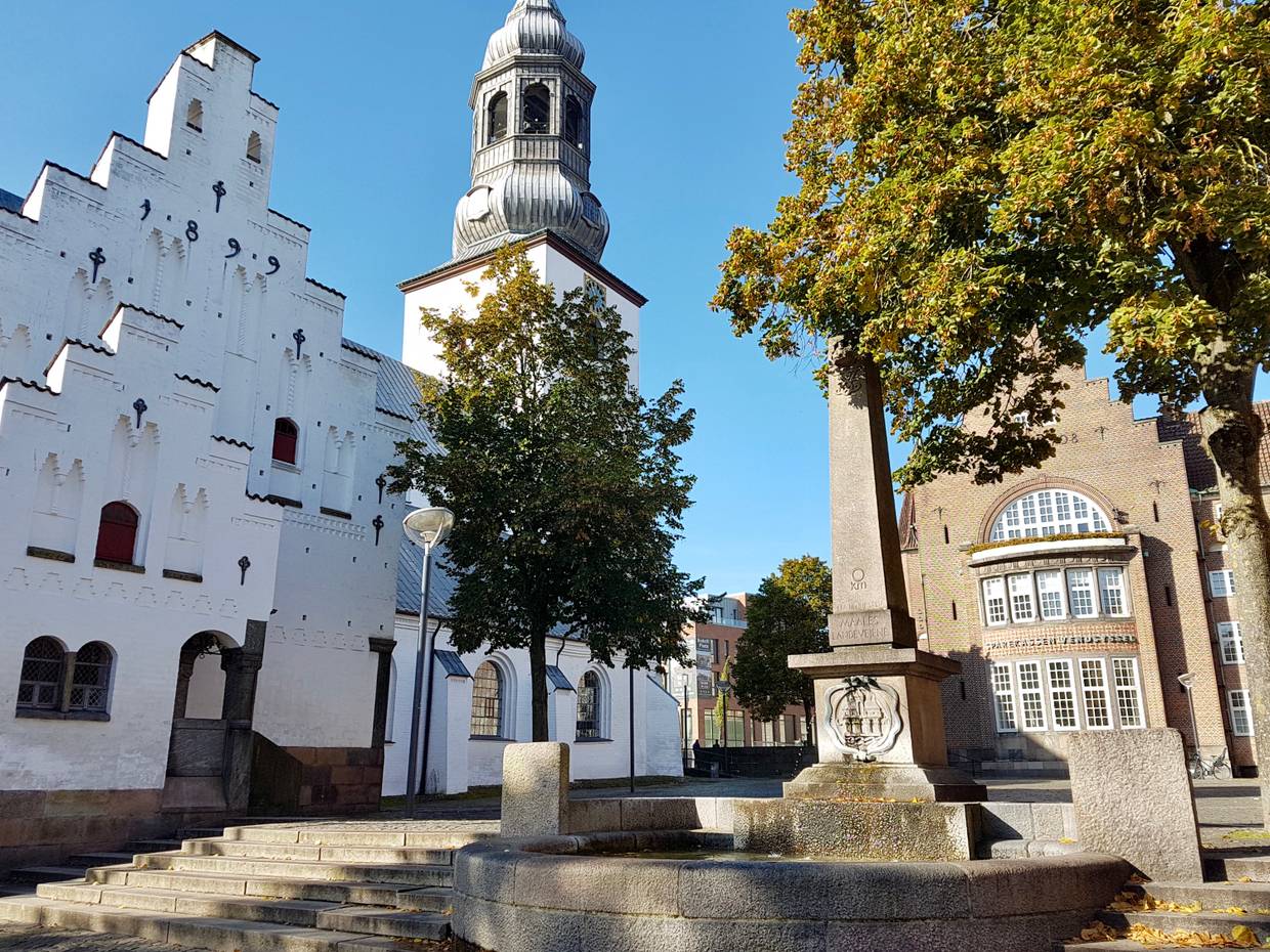 Obelist auf einem Sockel, dahinter die weiße Kirche