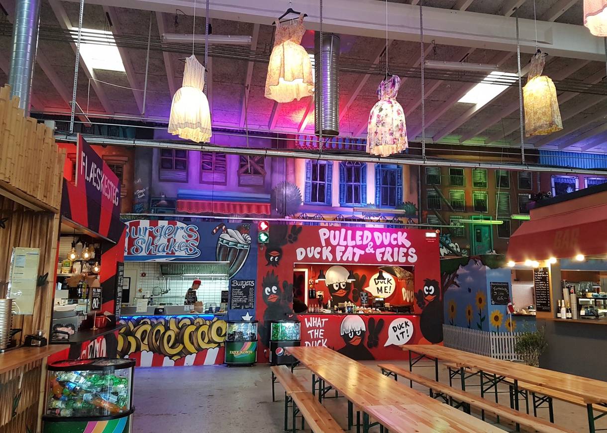 Streetfoodhalle mit bunt bemalten Ständen, Biergartengarnituren und bunten Kleidern als Lampenschirmen an der Decke