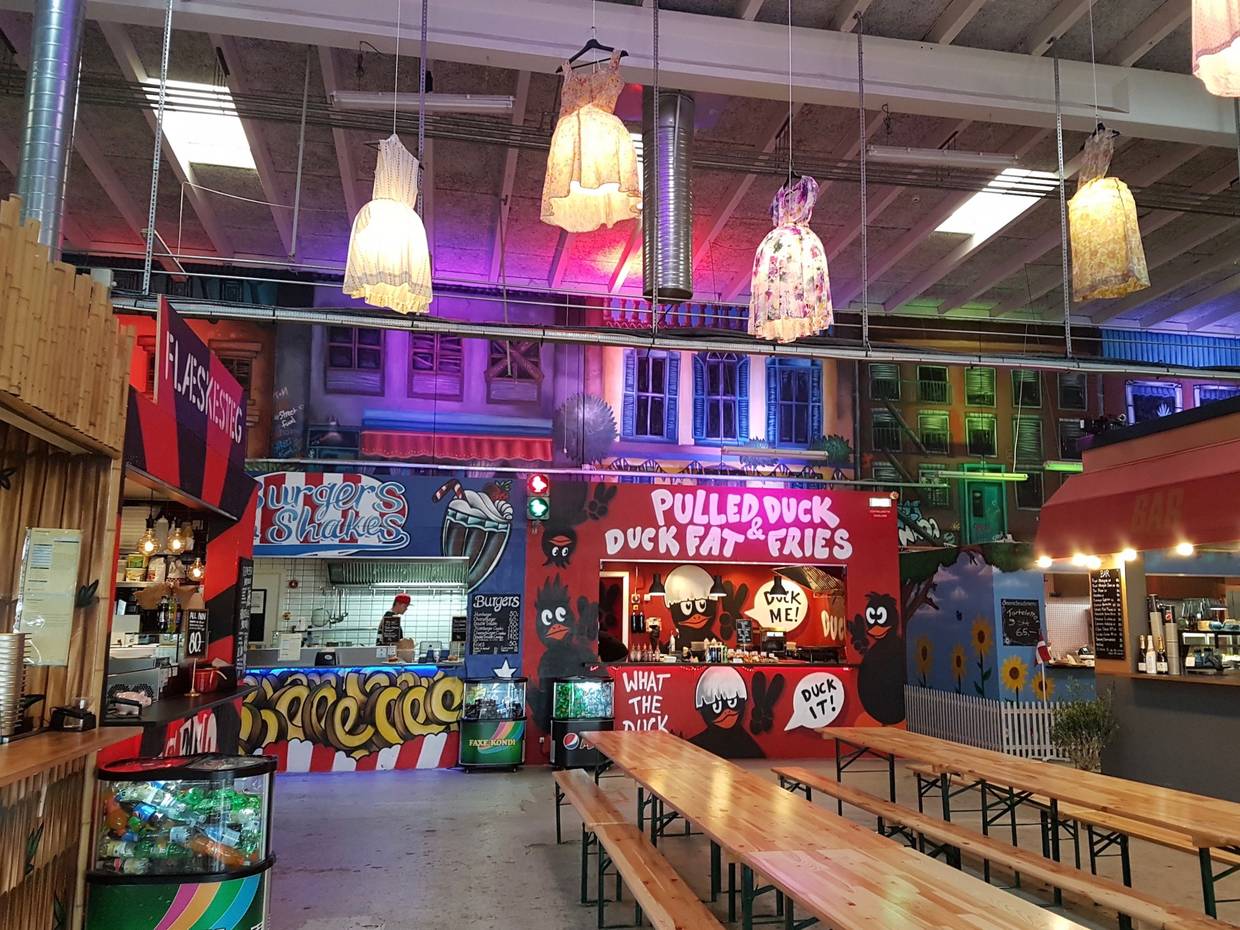Streetfoodhalle mit bunt bemalten Ständen, Biergartengarnituren und bunten Kleidern als Lampenschirmen an der Decke