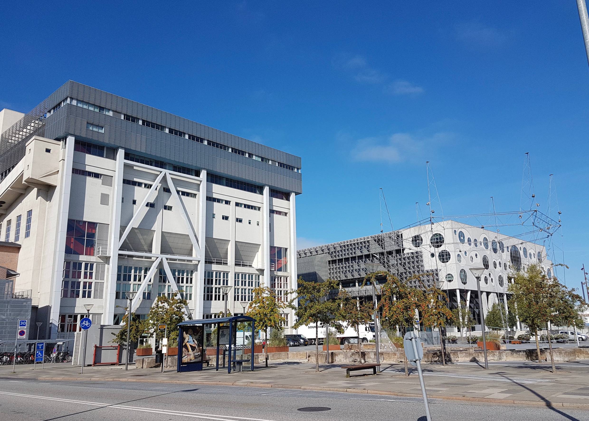Zwei moderne, weiß-graue Bauwerke, davor ein angelegter Platz mit jungen Bäumen