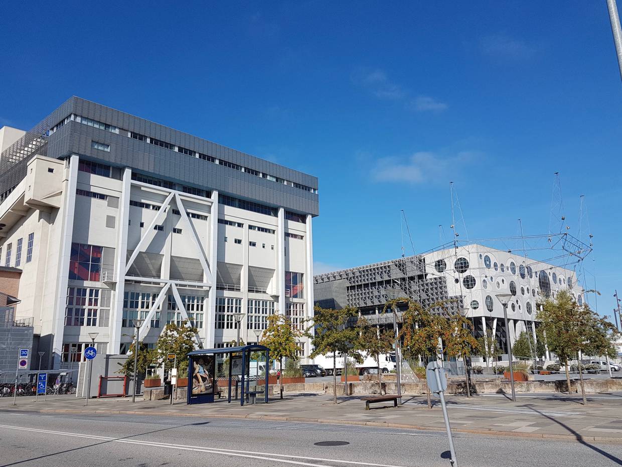 Zwei moderne, weiß-graue Bauwerke, davor ein angelegter Platz mit jungen Bäumen