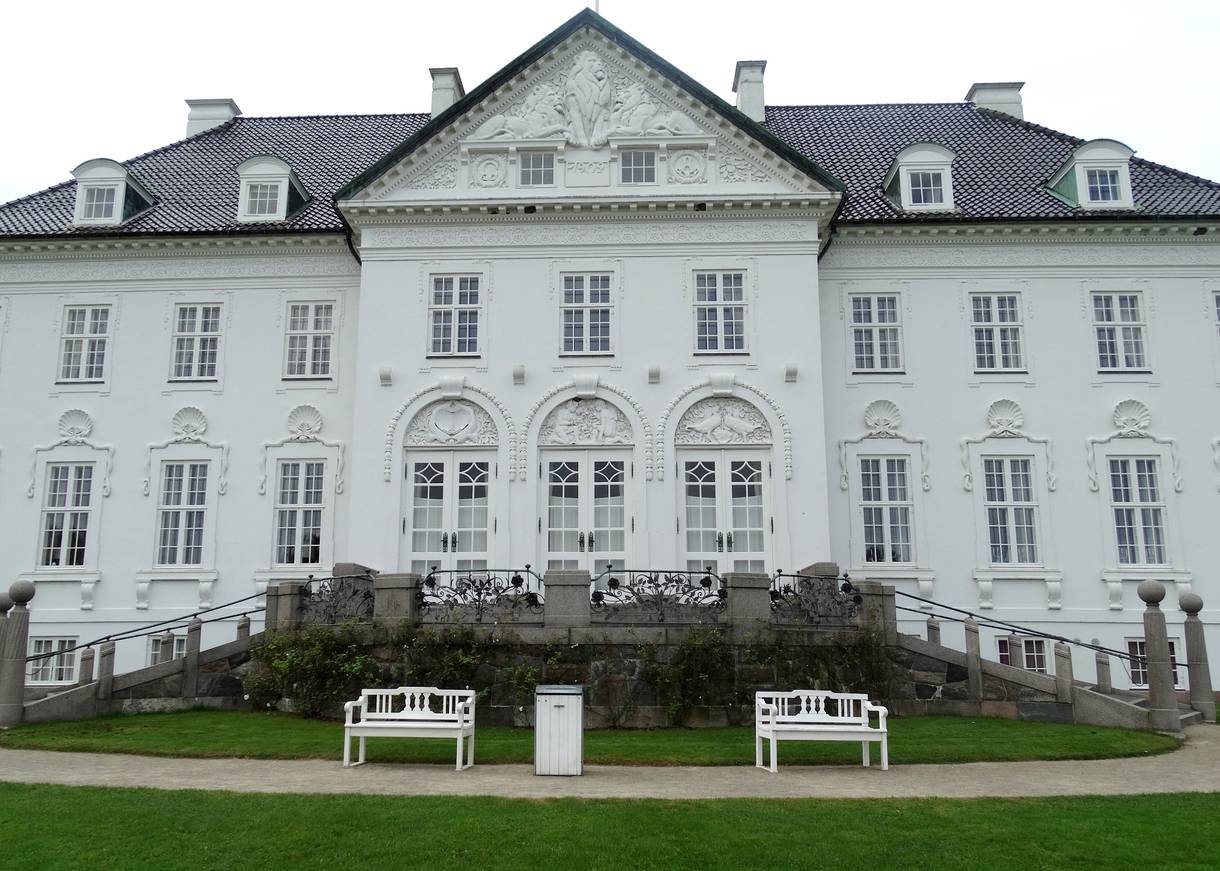 Weißes Herrenhäuser mit runden, reich verzierten Fensterbögen, davor zwei Bänke