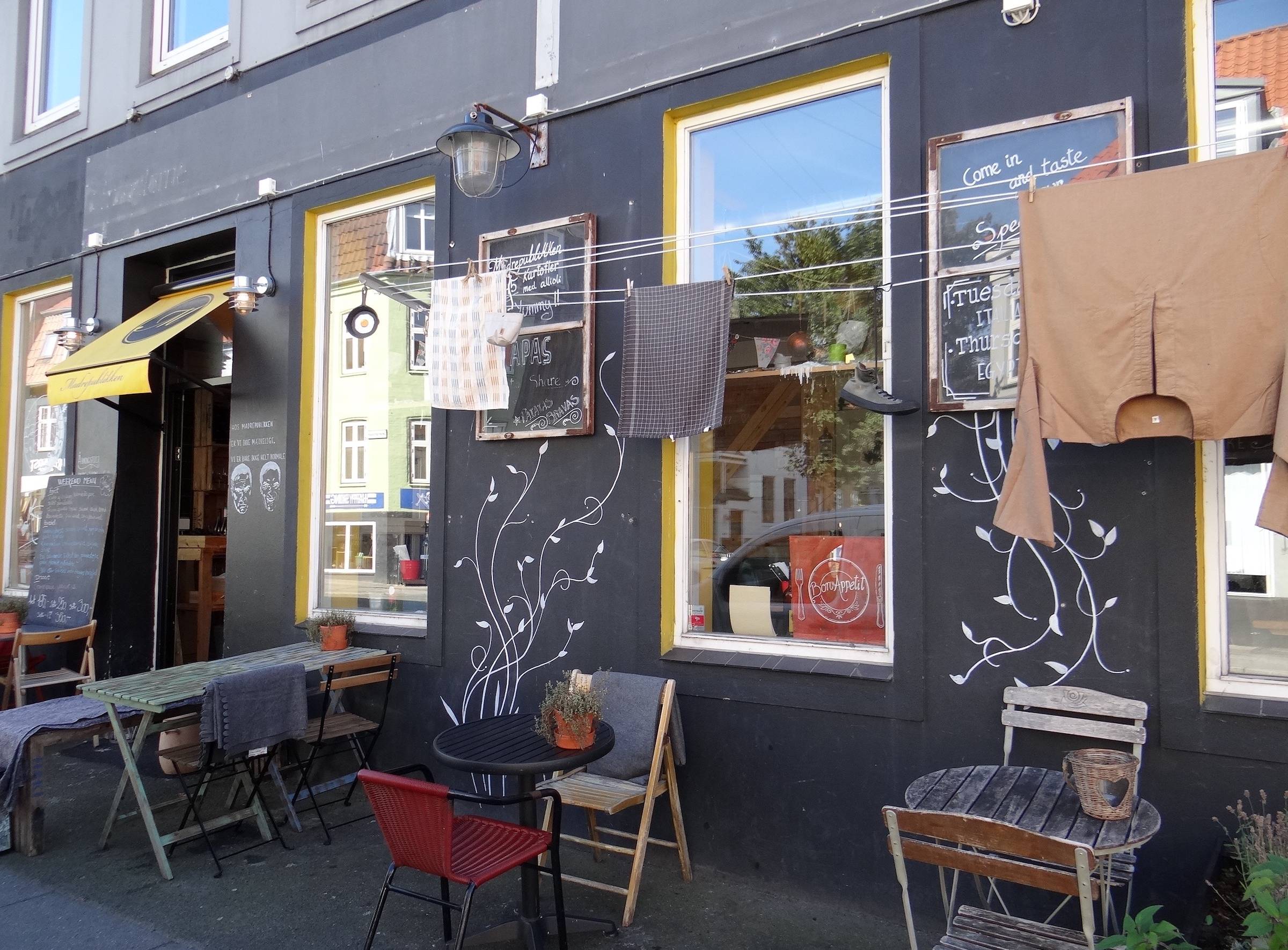 Fassade einer Pizzeria, die an eine Dorfszene in Italen erinnert, mit Wäsche an der Leine und unterschiedlichen, kleinen Tischen
