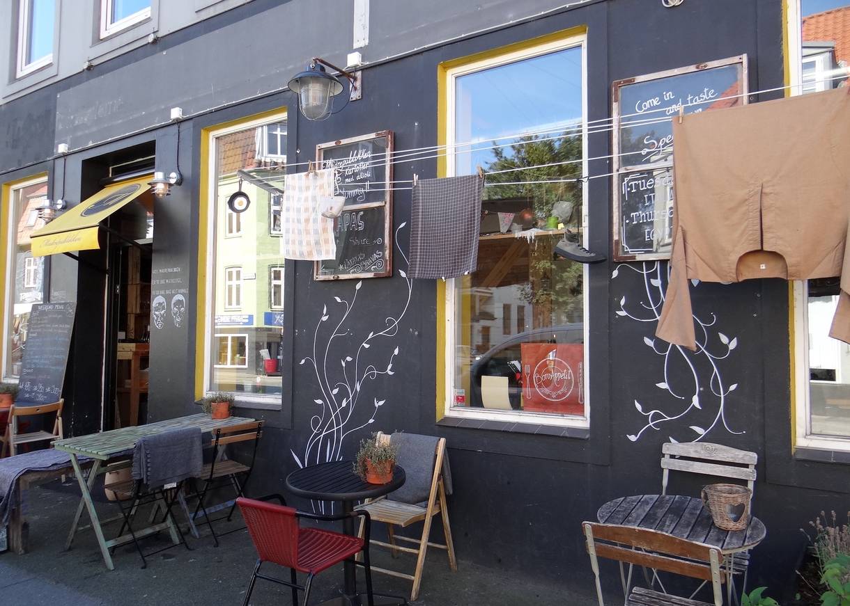 Fassade einer Pizzeria, die an eine Dorfszene in Italen erinnert, mit Wäsche an der Leine und unterschiedlichen, kleinen Tischen