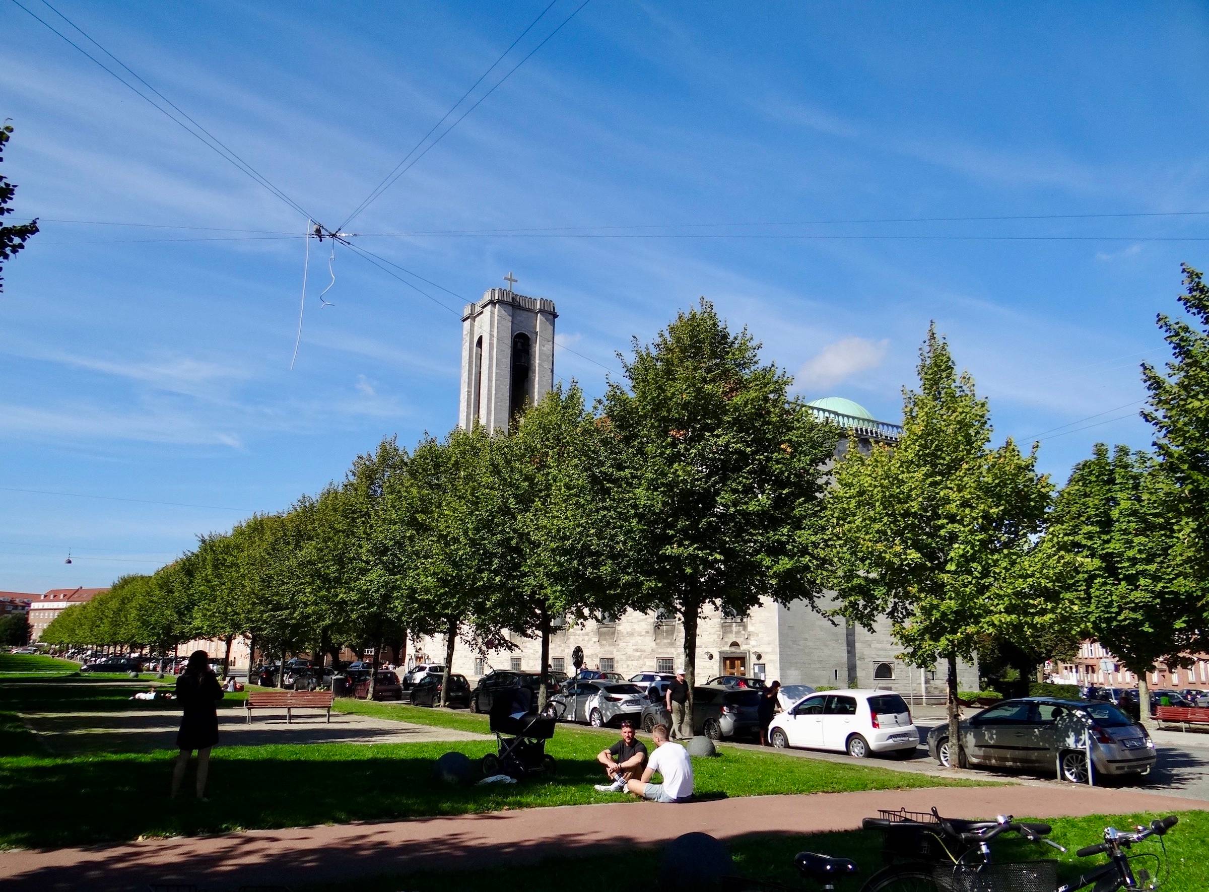 Allee mit kleinen Bäumen, dazwischen sitzen Leute auf dem Rasen, im Hintergrund eine moderne Kirche