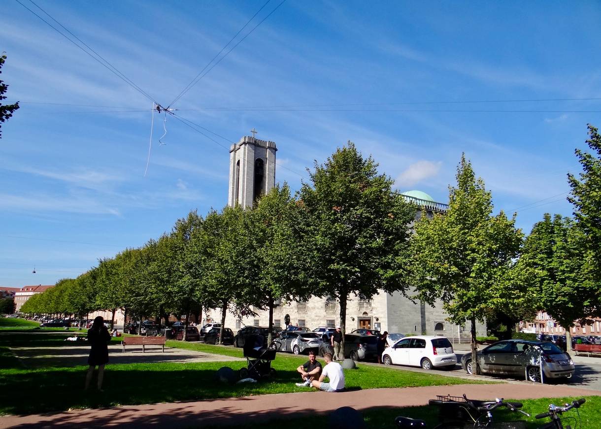 Allee mit kleinen Bäumen, dazwischen sitzen Leute auf dem Rasen, im Hintergrund eine moderne Kirche