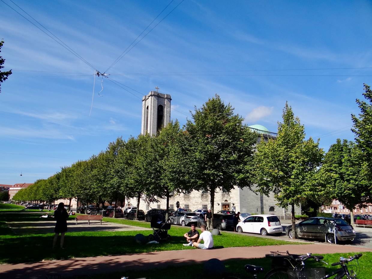 Allee mit kleinen Bäumen, dazwischen sitzen Leute auf dem Rasen, im Hintergrund eine moderne Kirche