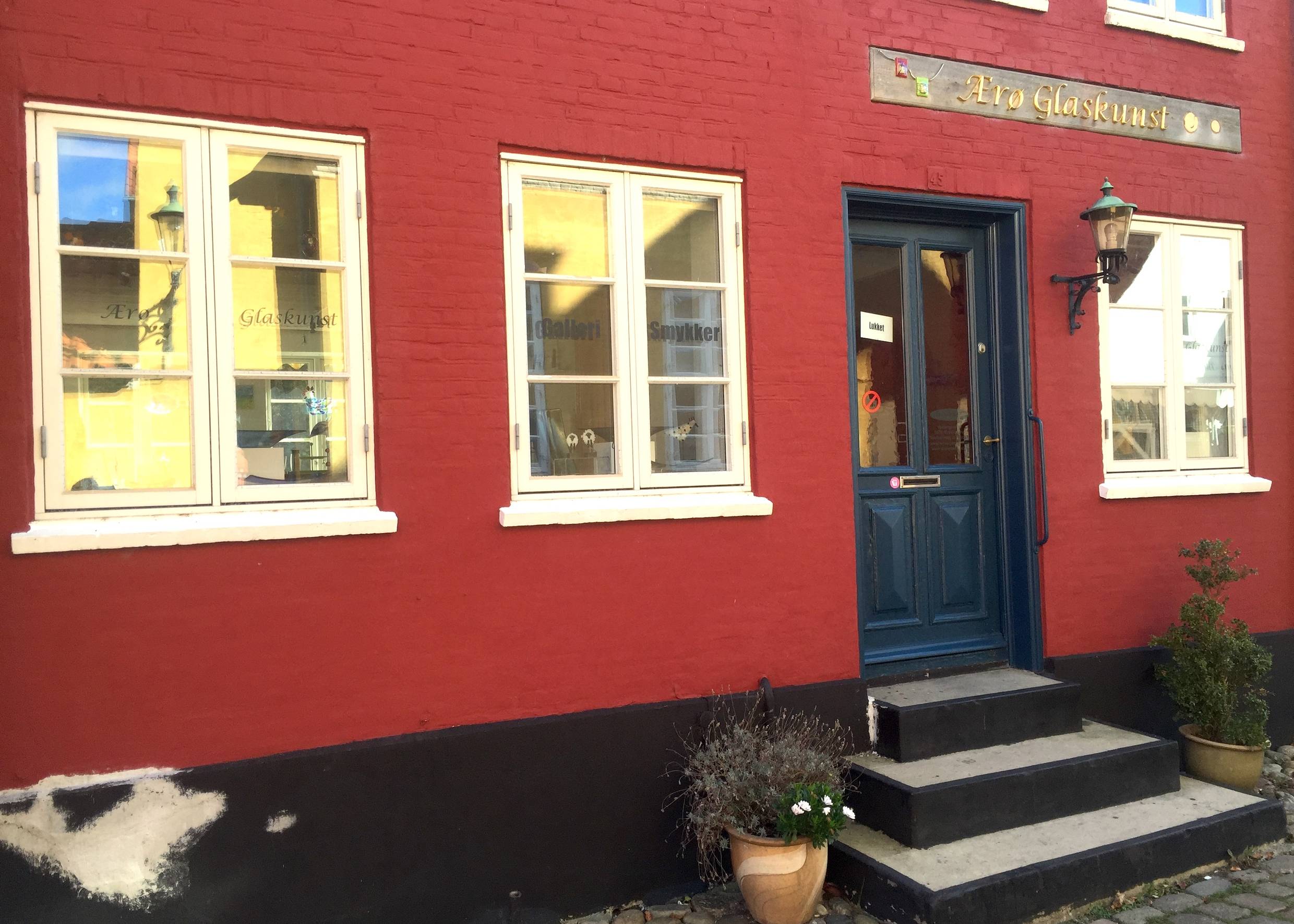 Ein rotes Wohnhaus mit weißen Fenstern, dunkelblaue Tür, Eingang zu einer Galerie