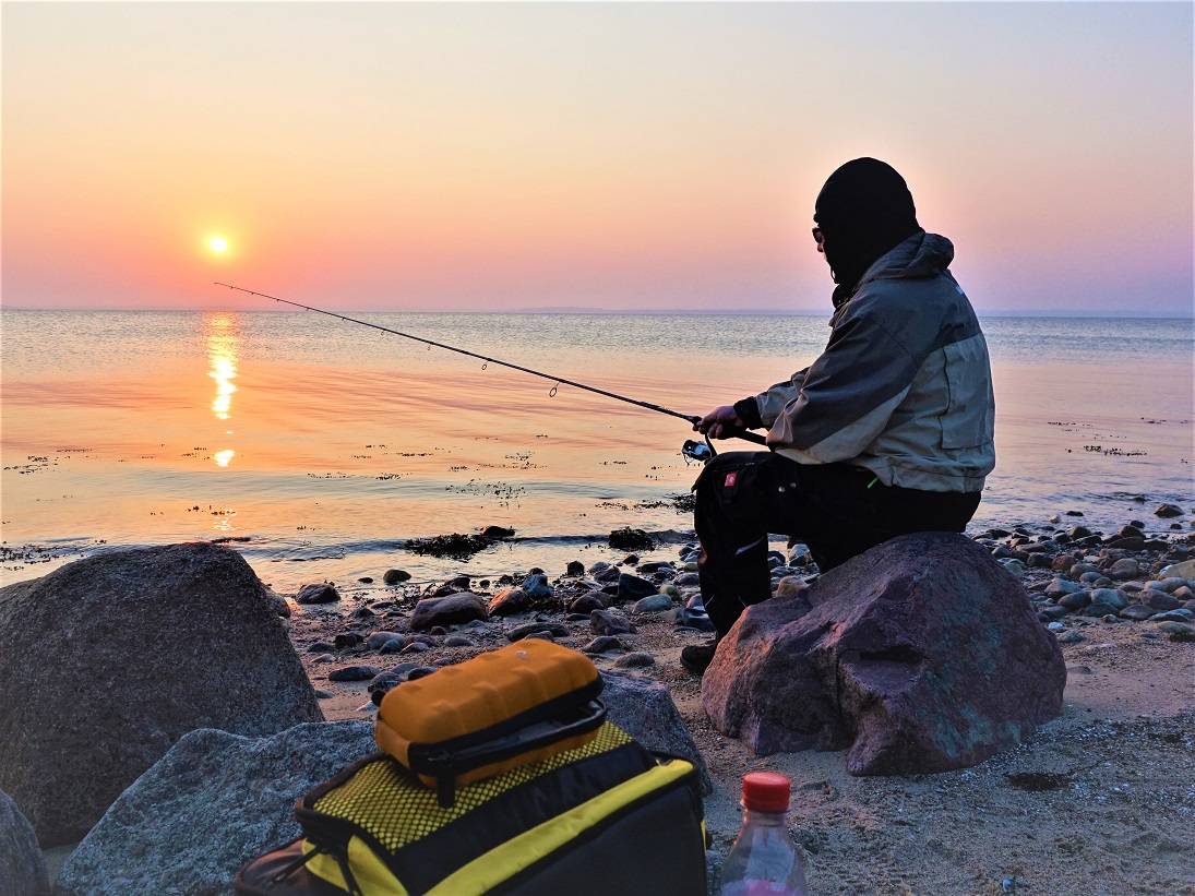 Angler auf einem Stein am Strand vor aufgehender Sonne