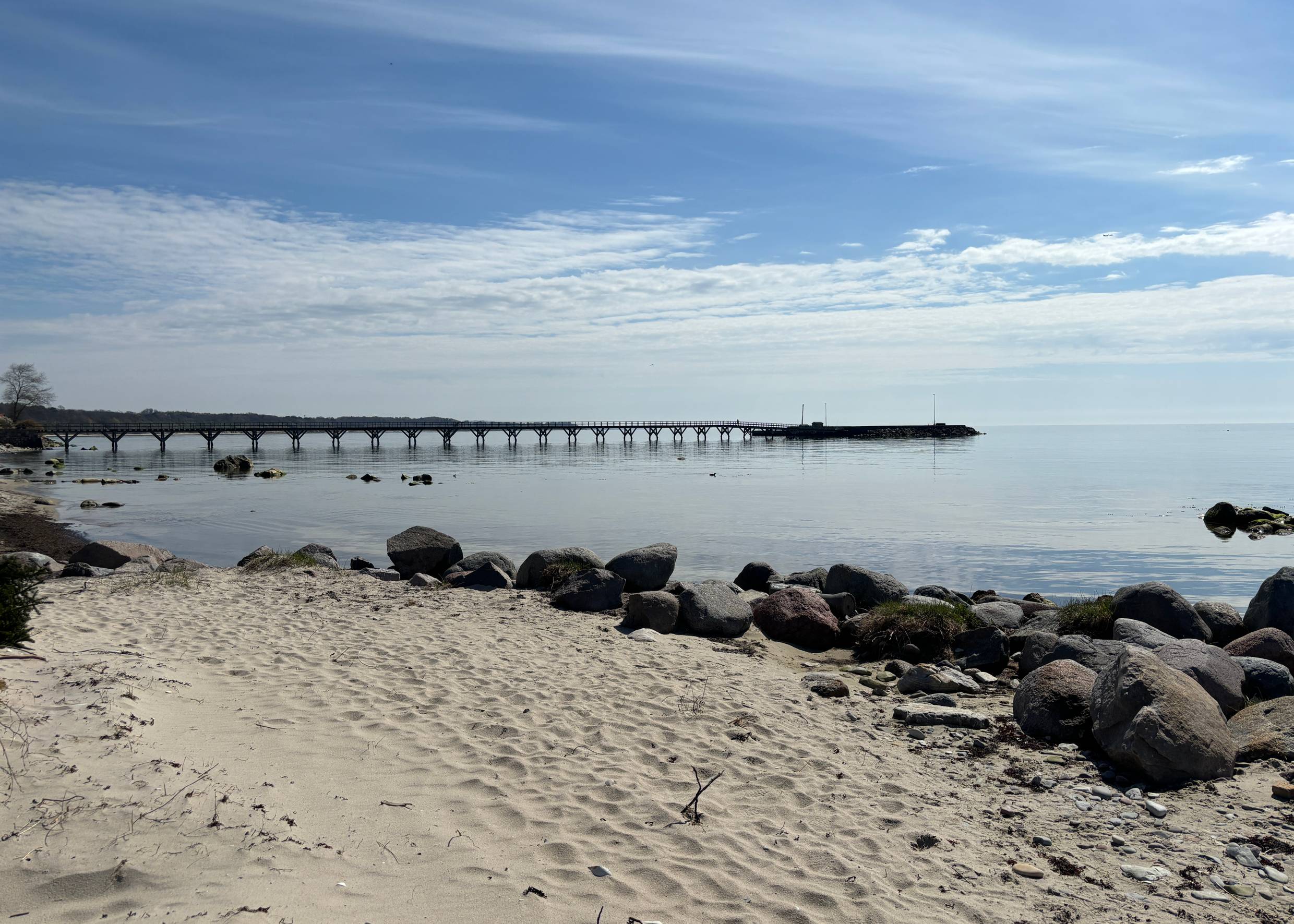 Ein langer Steg ragt ins Meer, vorne im Bild en schmaler Sandstrand