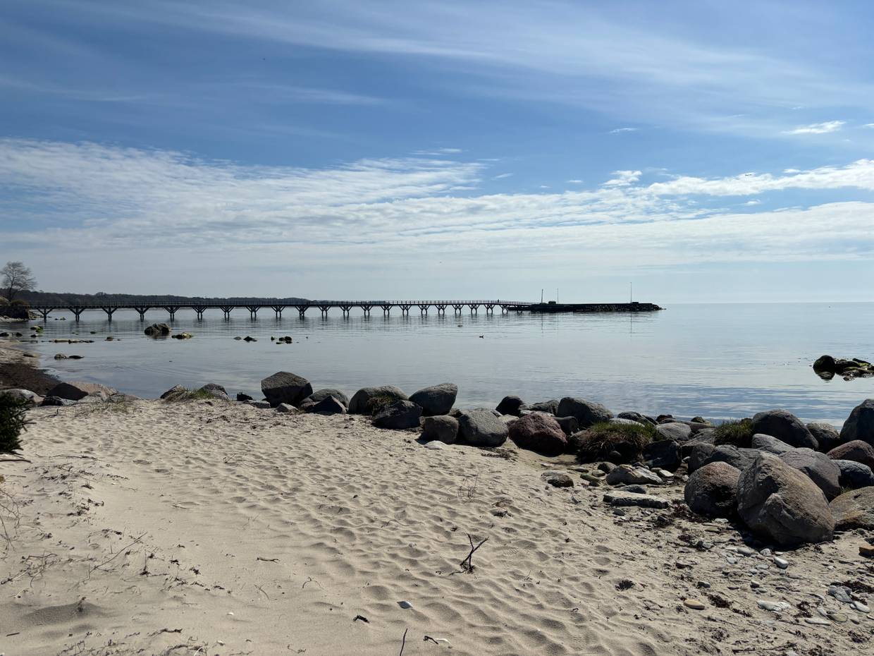 Ein langer Steg ragt ins Meer, vorne im Bild en schmaler Sandstrand
