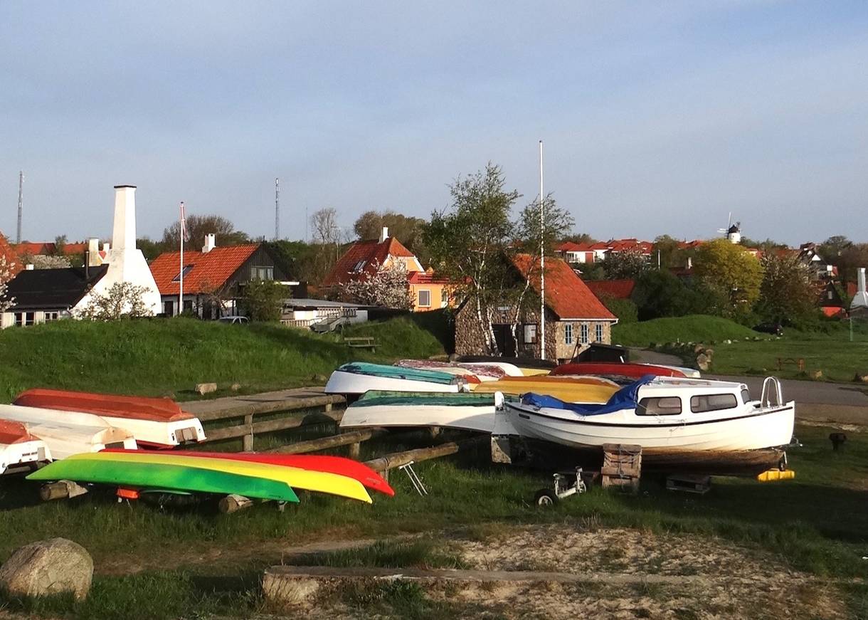 Boote am Strand, dahinter ein Dorf mit roten Dächern und weißen Schornsteinen