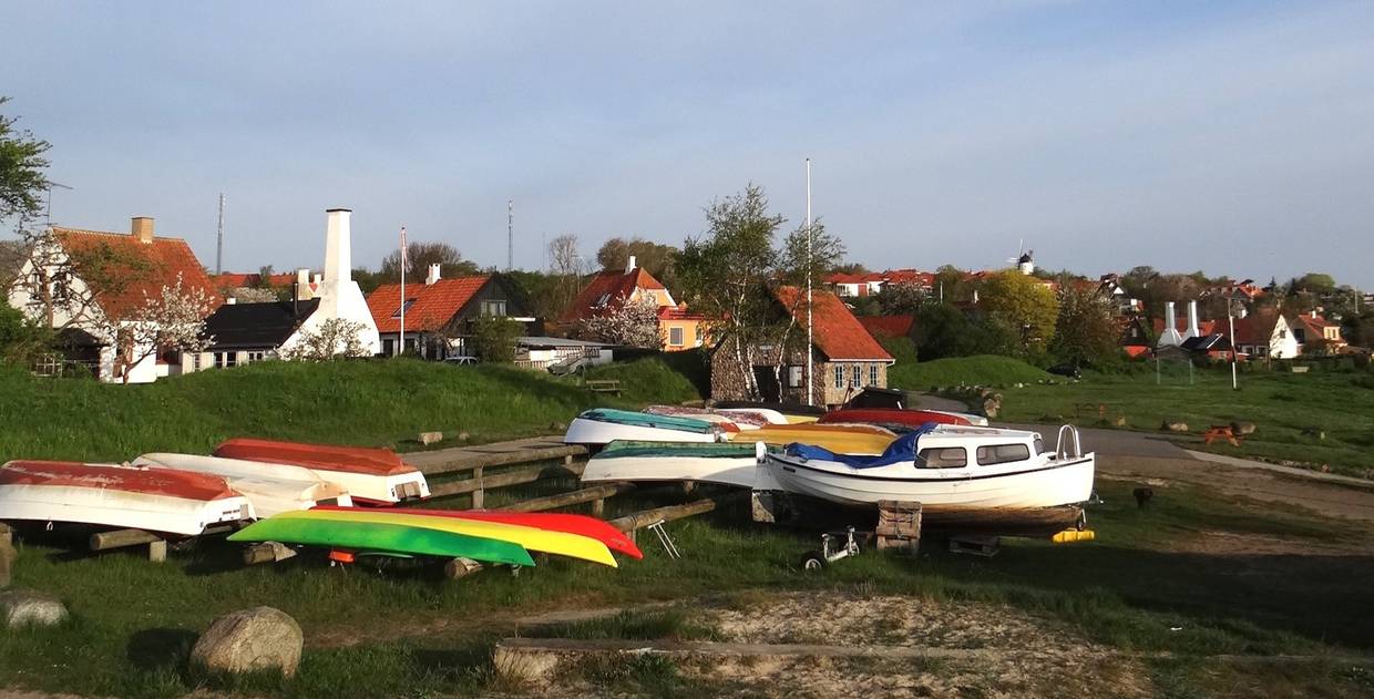 Boote am Strand, dahinter ein Dorf mit roten Dächern und weißen Schornsteinen