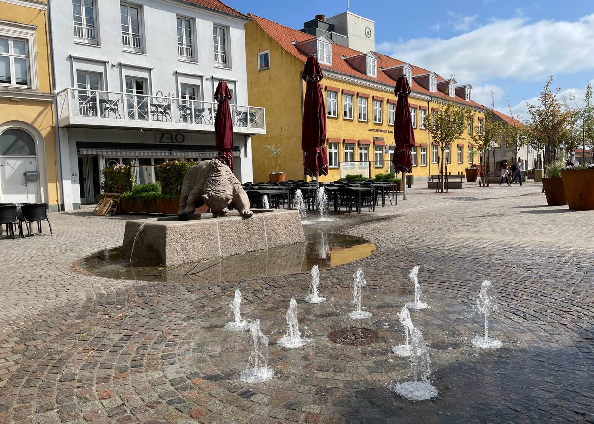 Fußgängerzone mit kleinen älteren Häusern, in der Mitte kleine Wasserfontänen