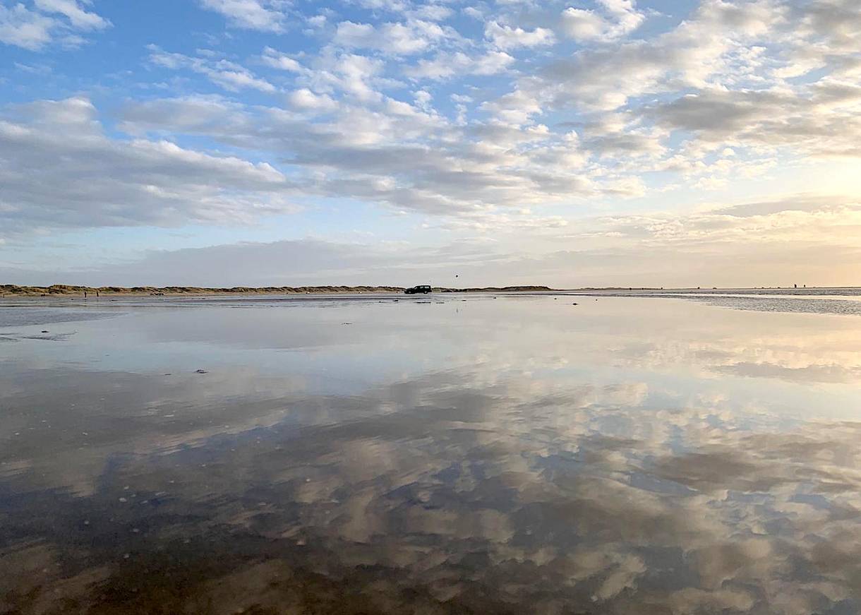 Spiegelnde Wasserfläche auf dem Strand, dahinter ein Auto