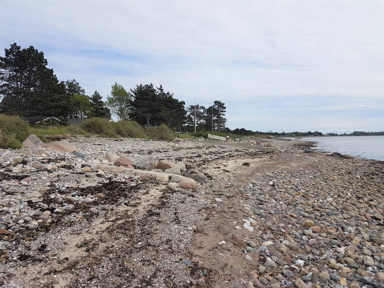 Sehr steiniger Strand, dahinter Ferienhäuser zwischen den Bäuemen