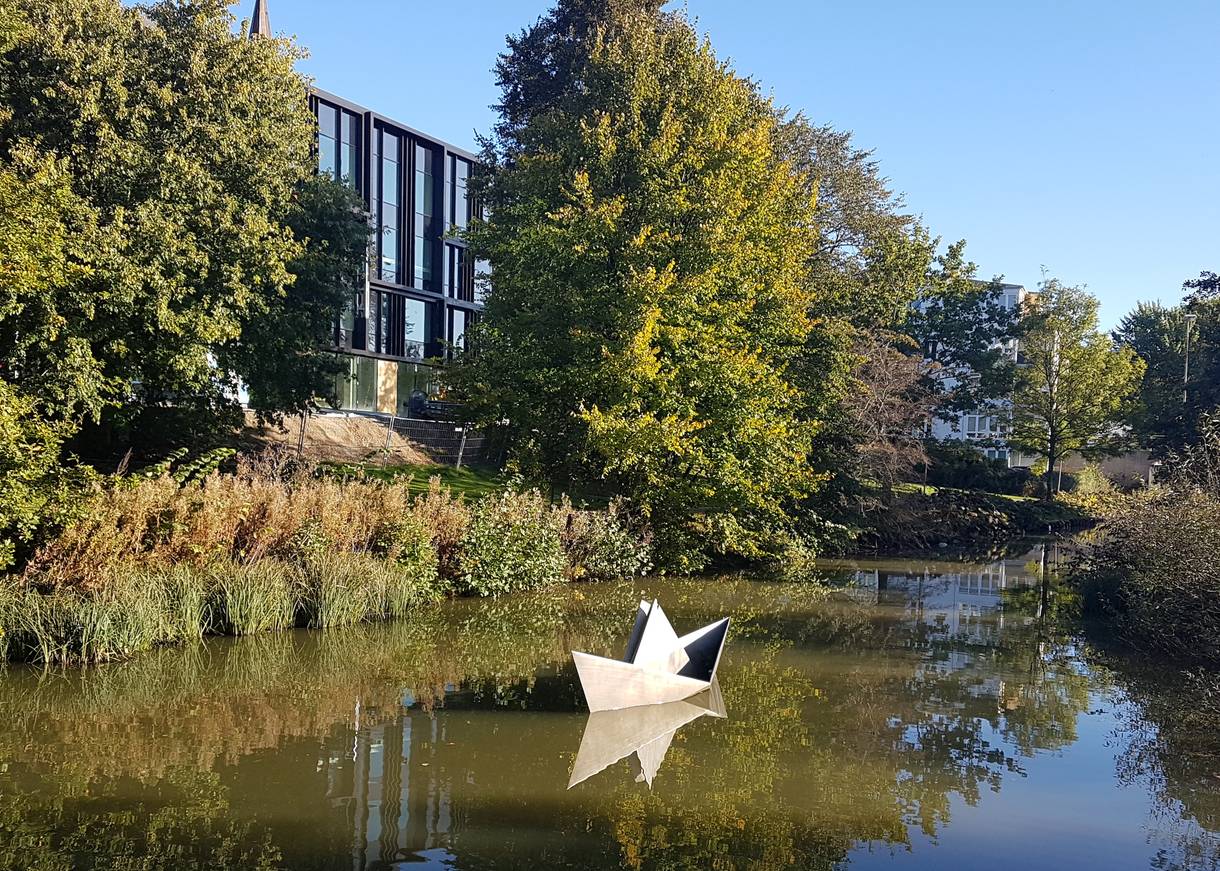Skulptur eines Papierschiffes auf einem Bach