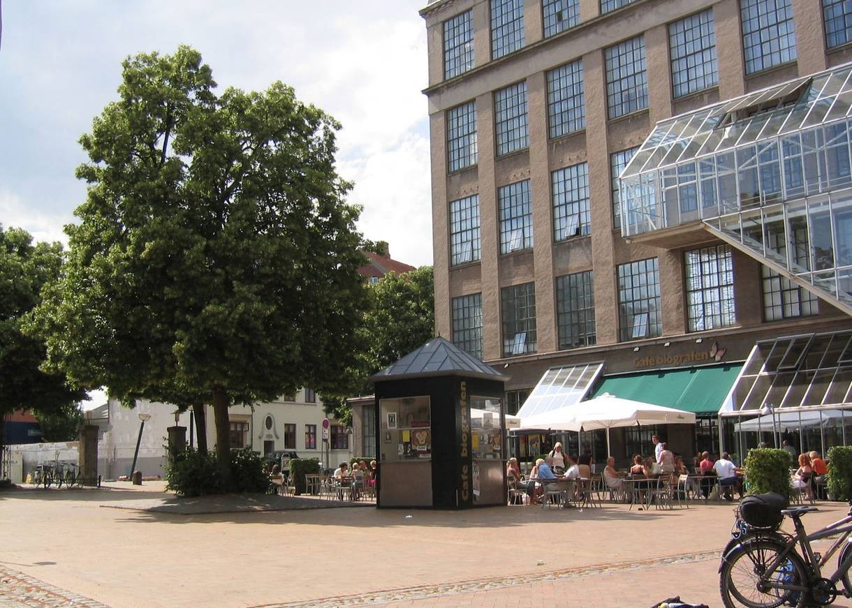 Platz vor einem ehemaligen Fabrikgebäude mit einem Baum, dahinter ein Restaurant
