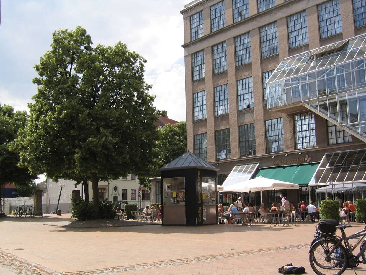 Platz vor einem ehemaligen Fabrikgebäude mit einem Baum, dahinter ein Restaurant