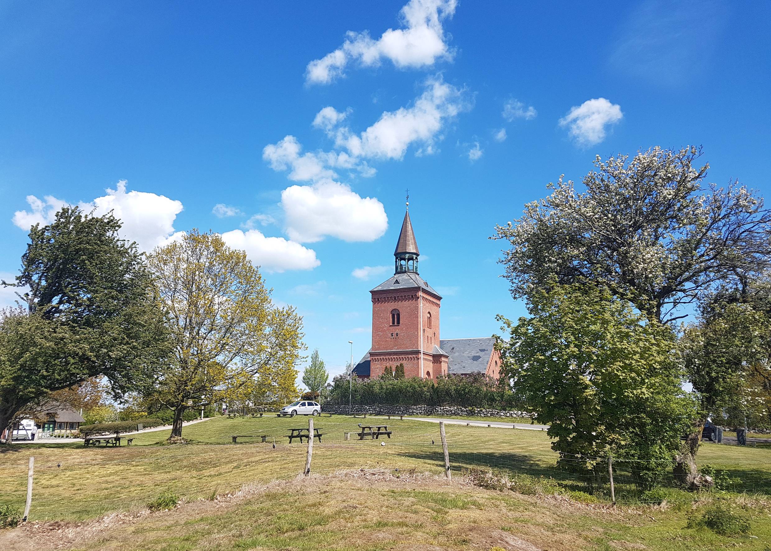 Blick über ein Feld mit Picknickbänken, einige Bäume und eine rote Backsteinkirche auf Hügelchen