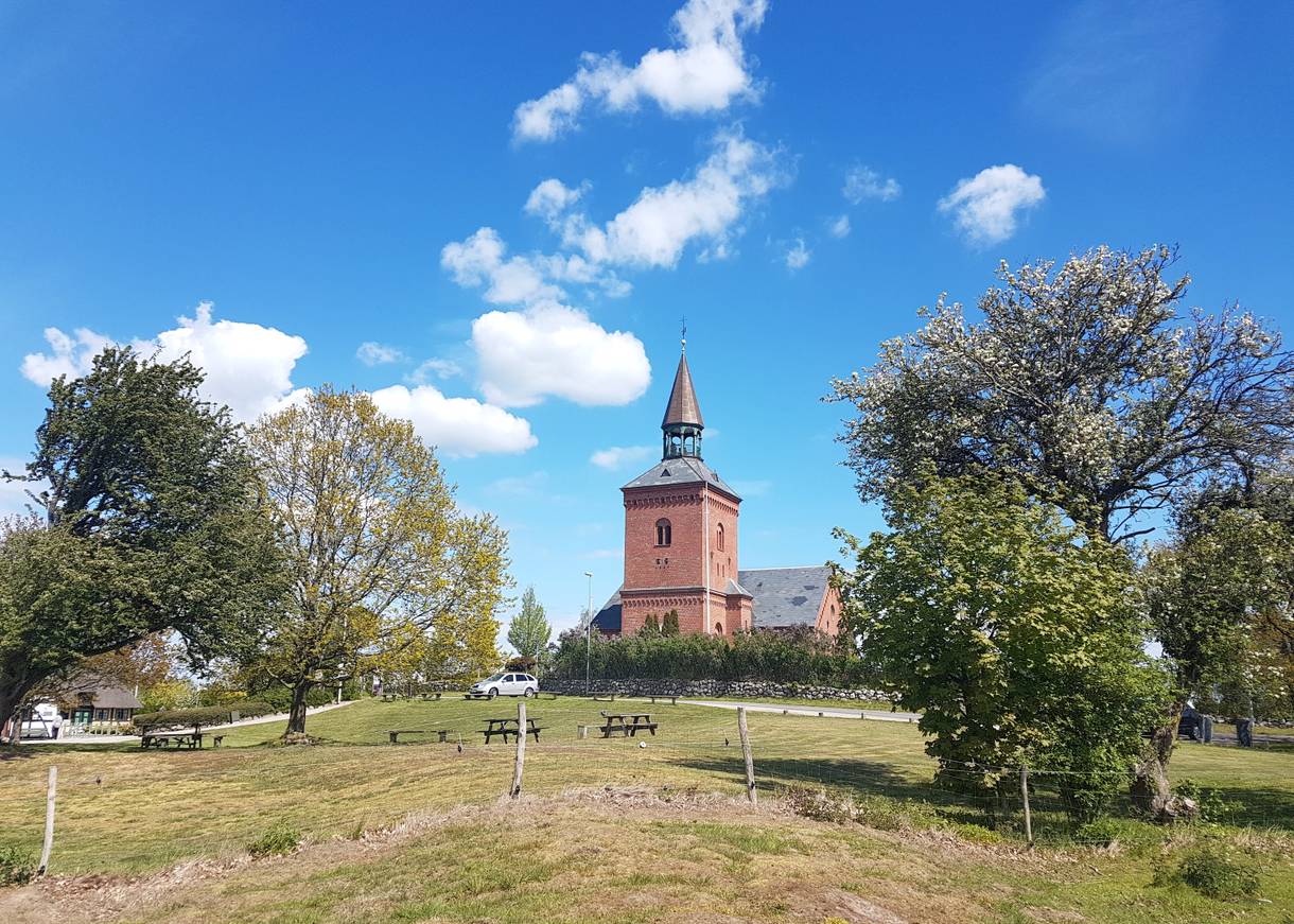 Blick über ein Feld mit Picknickbänken, einige Bäume und eine rote Backsteinkirche auf Hügelchen