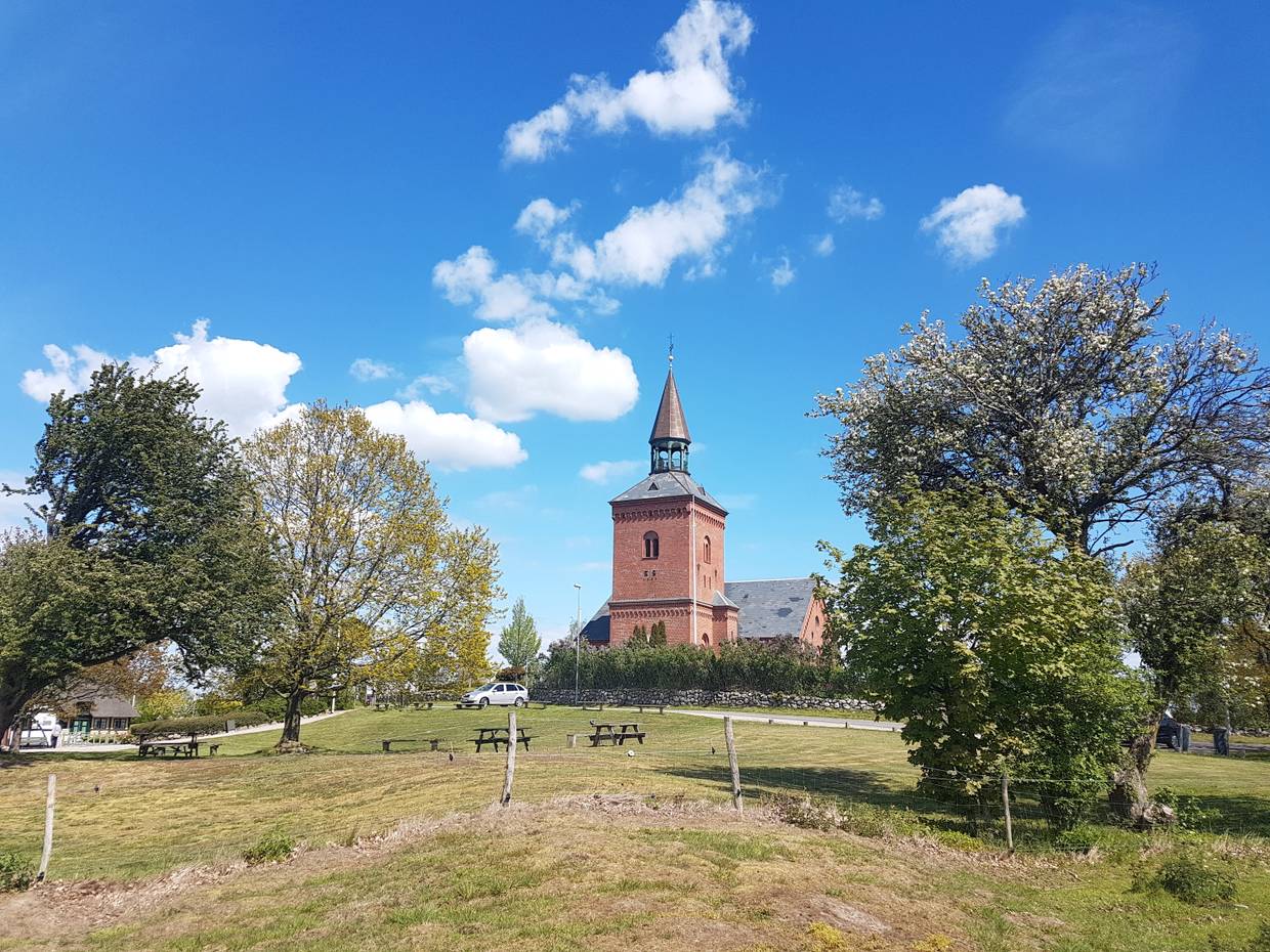 Blick über ein Feld mit Picknickbänken, einige Bäume und eine rote Backsteinkirche auf Hügelchen