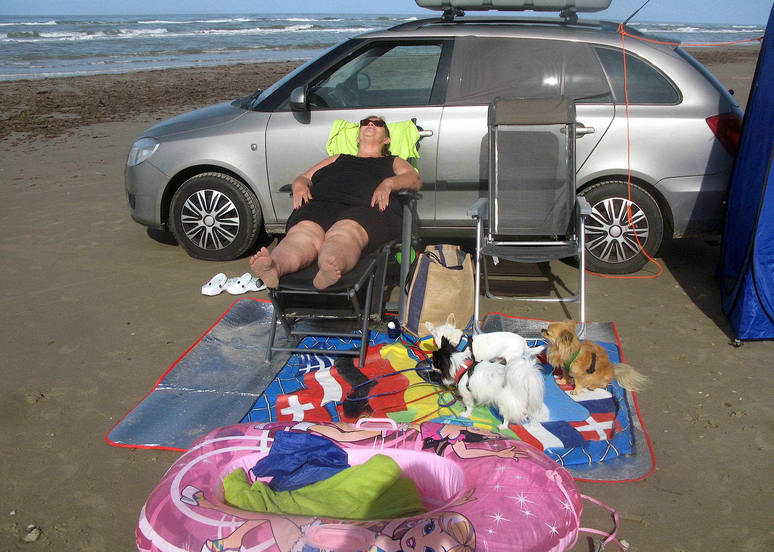 Frau auf einem Liegestuhl am Strand, dahinter das Auto, davor allerlei Strandzubehör