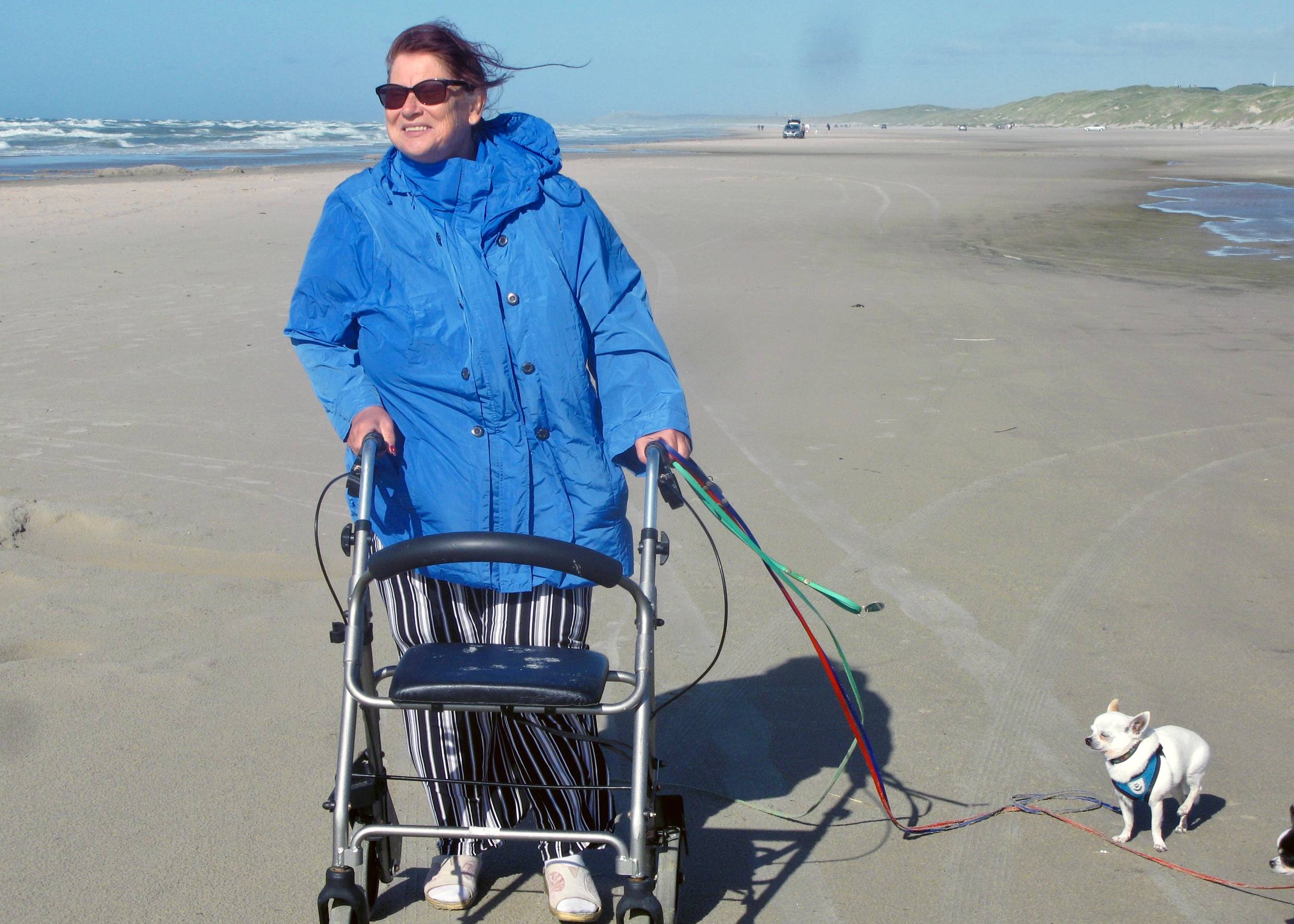 Frau mit Rollator und einem kleinen Hund an der Leine auf einem breiten Sandstrand