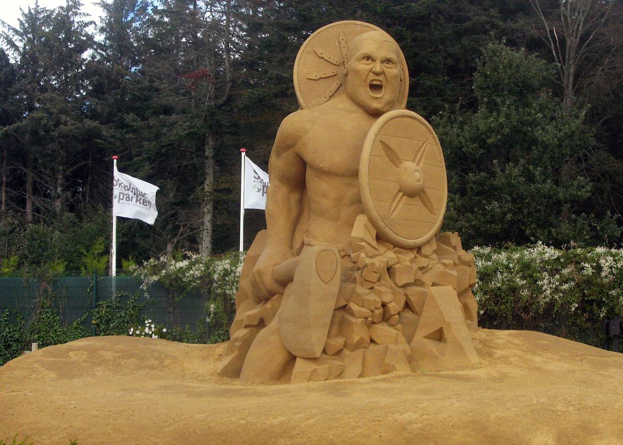 Figur aus Sand, im Hintergrund Flaggen des Skulpturenparks