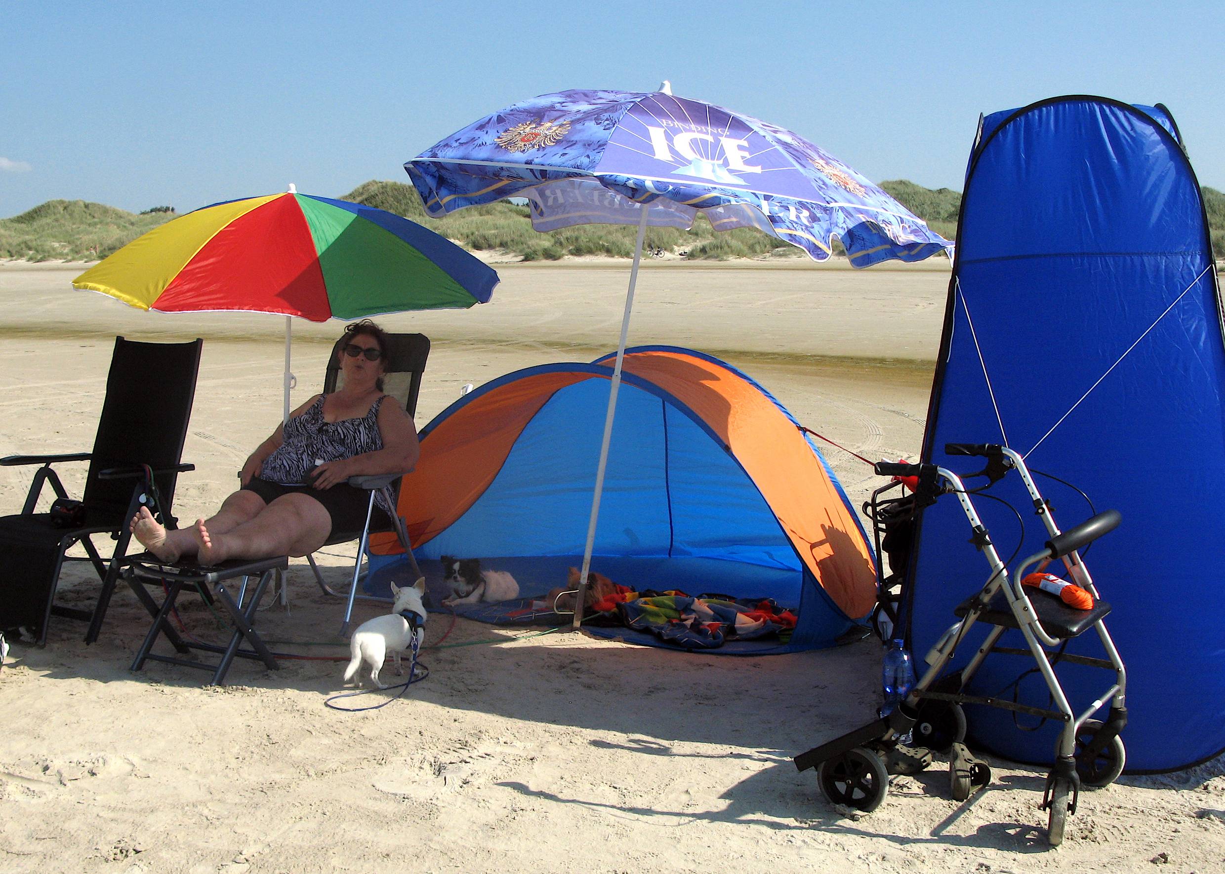 Frau auf einem Liegestuhl am Strand, zwei Sonnenschirme und ein blaues Zelt daneben