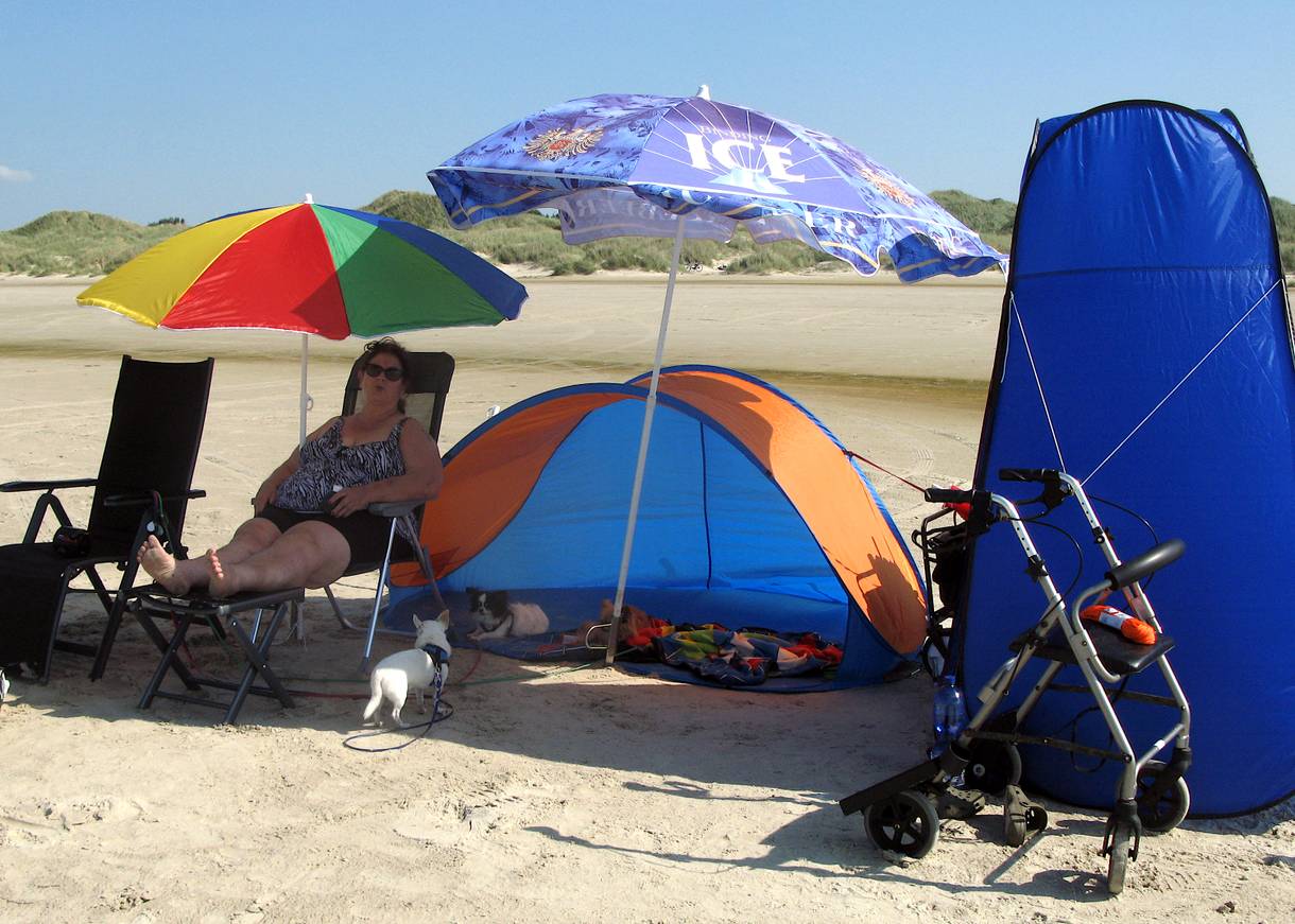 Frau auf einem Liegestuhl am Strand, zwei Sonnenschirme und ein blaues Zelt daneben