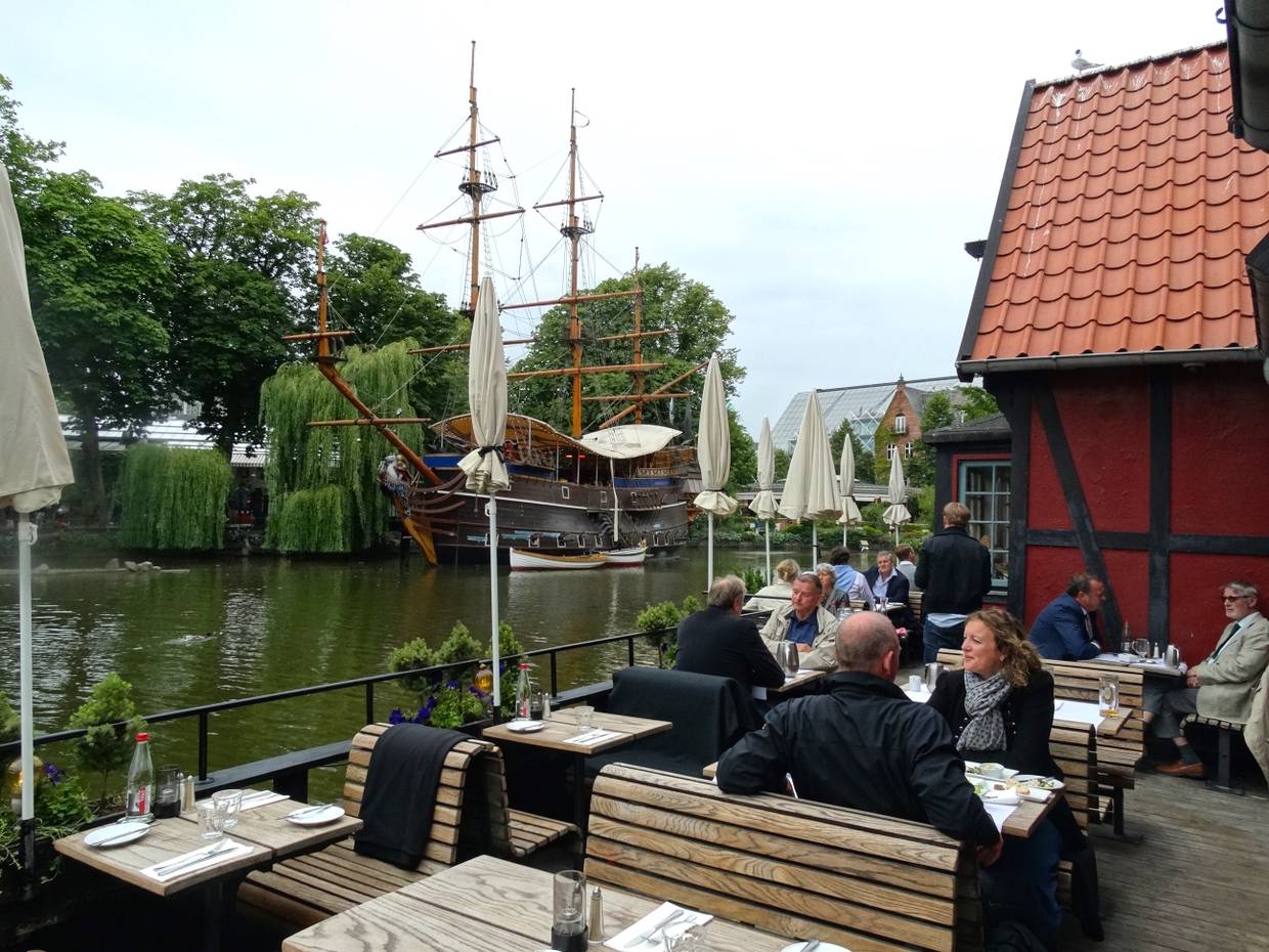 Restaurantterrasse an einem See, im Hintergrund ein Segelschiff