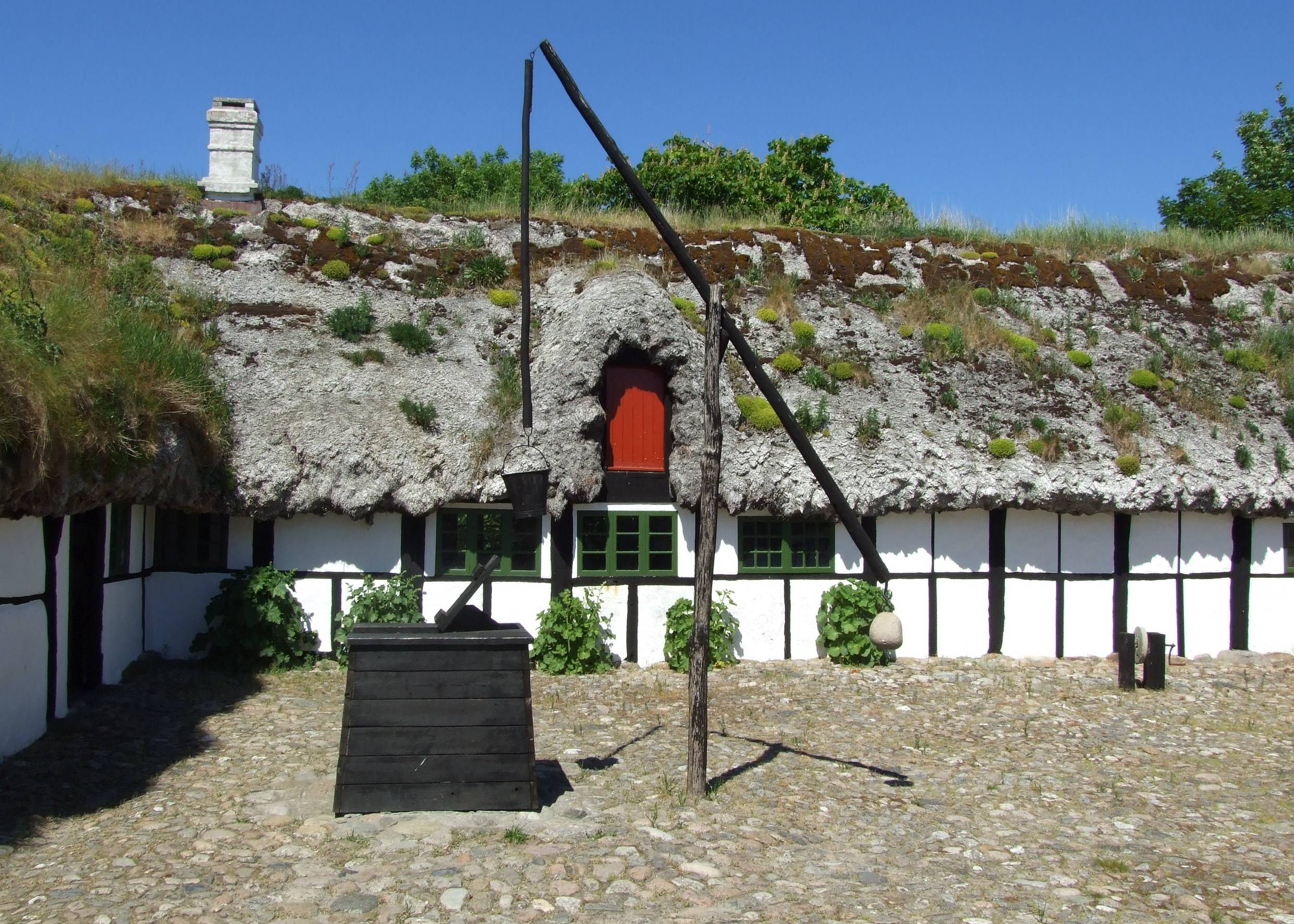 Innenhof des Læsø Museum, ein flaches weißes Bauernhaus mit einem Dach aus Seegras, davor ein schwarzer Ziehbrunnen
