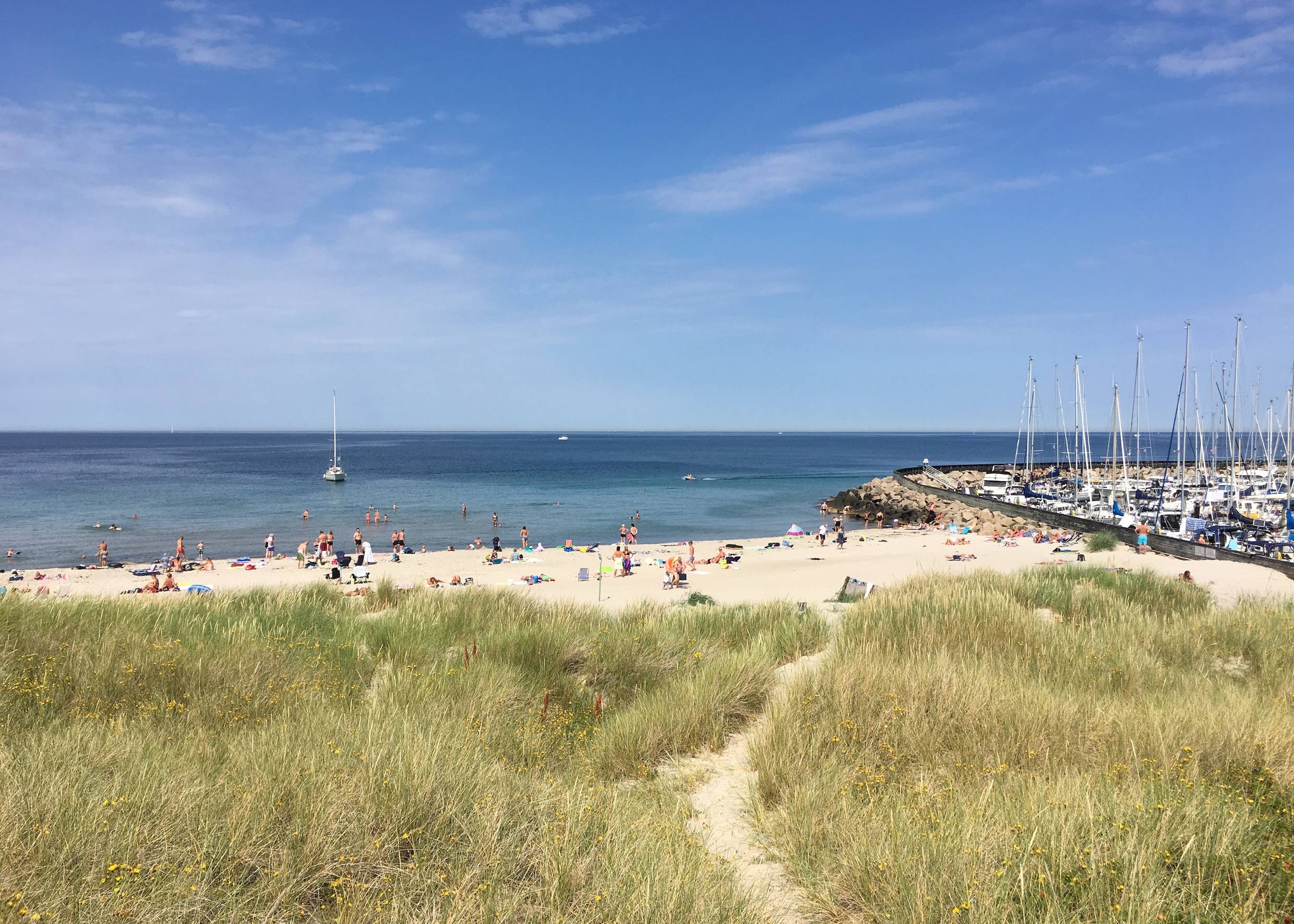 Sandstrand mit Badegäste, nebenan ein Jachthafen