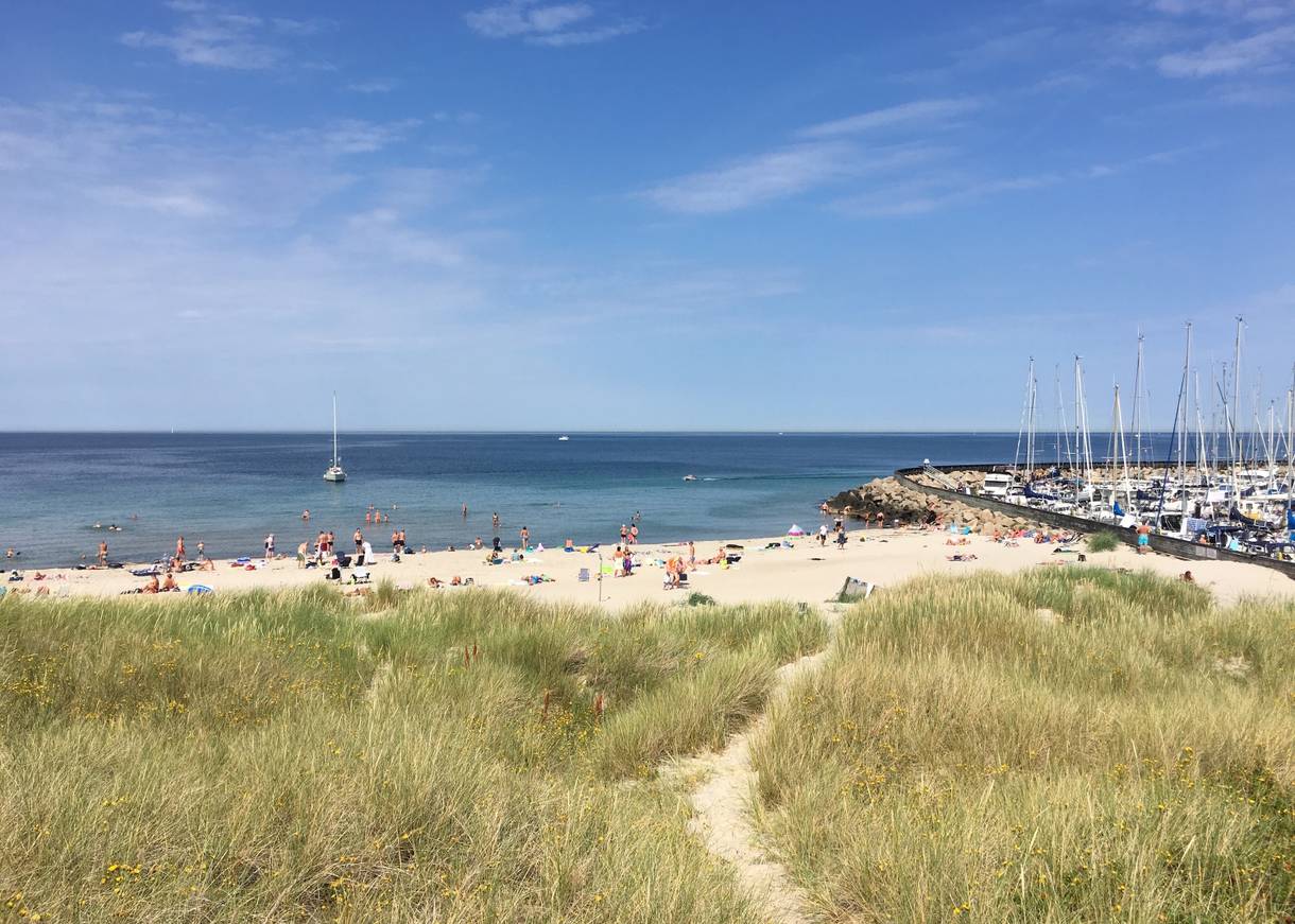 Sandstrand mit Badegäste, nebenan ein Jachthafen
