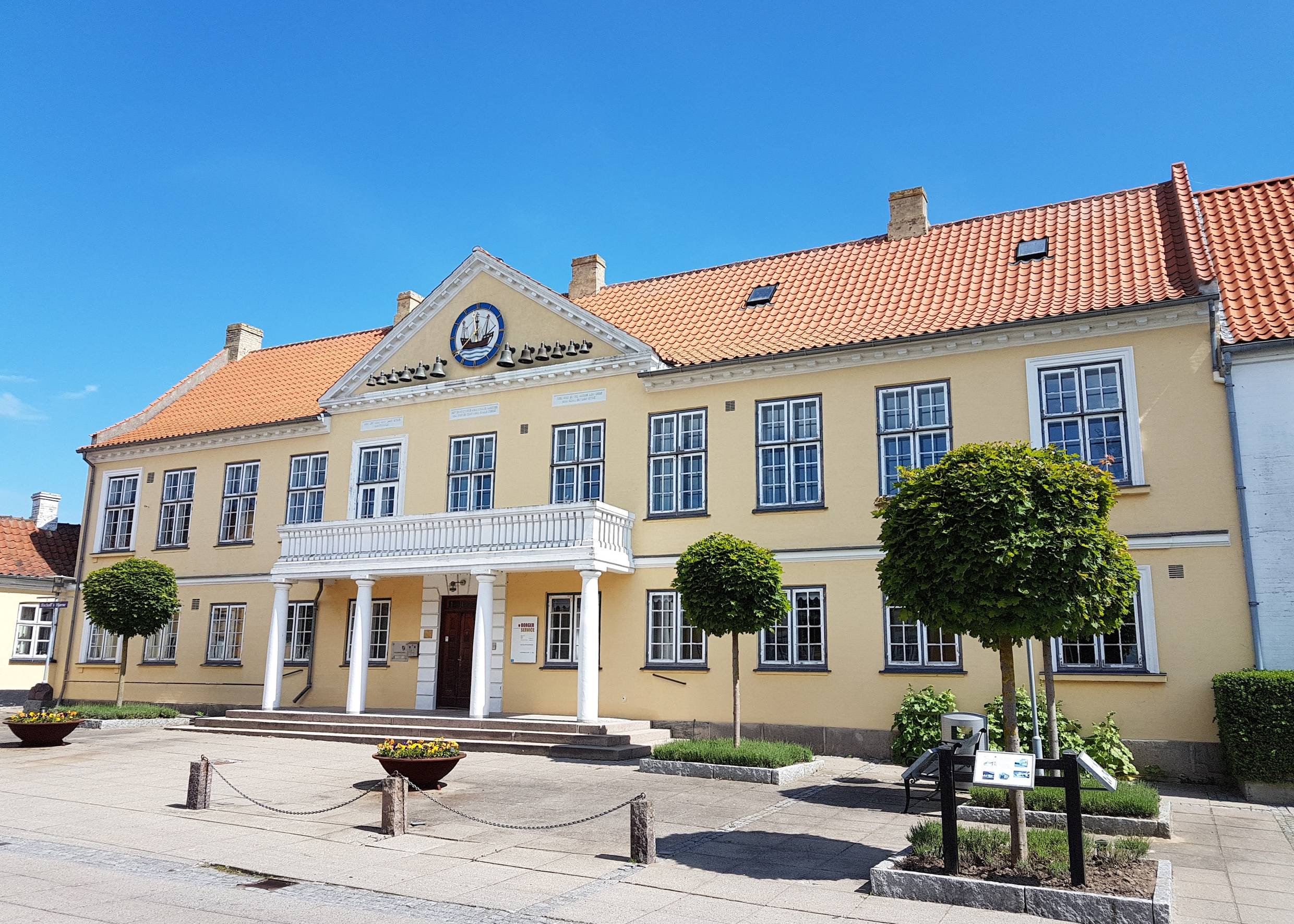 Hellgelbes Gebäude mit einem Eingang aus weißen Säulen, davor Blumenkübel