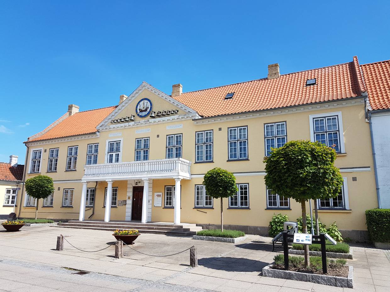 Hellgelbes Gebäude mit einem Eingang aus weißen Säulen, davor Blumenkübel