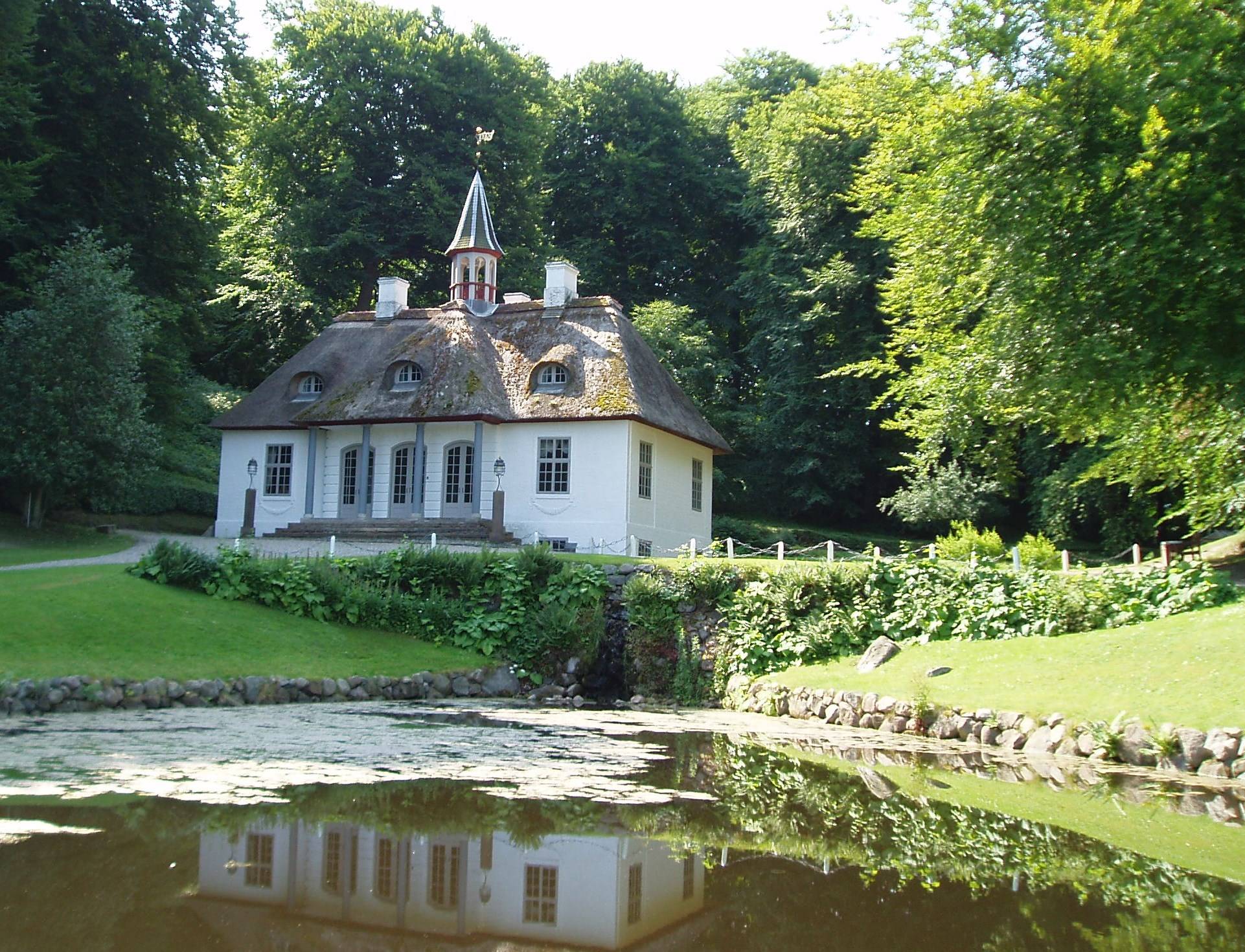 Blick auf ein weißes Schlössschen umgeben von Bäumen, davor ein See
