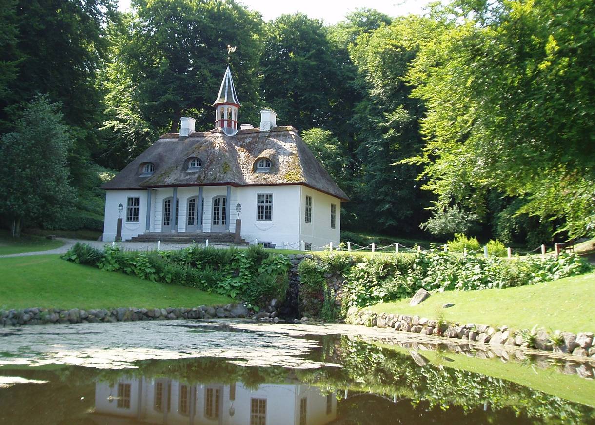 Blick auf ein weißes Schlössschen umgeben von Bäumen, davor ein See