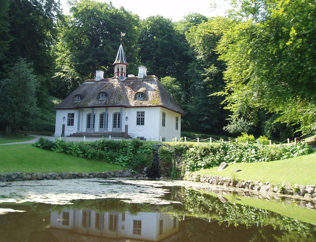 Blick auf ein weißes Schlössschen umgeben von Bäumen, davor ein See