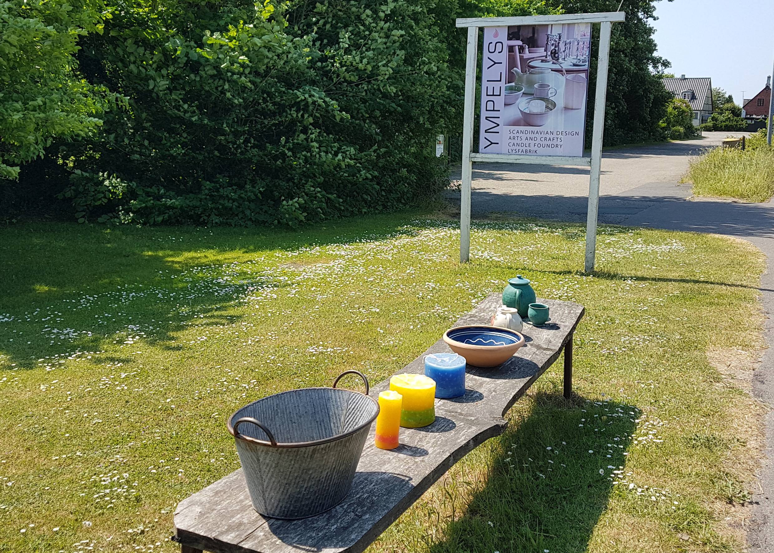 Holzbank am Wegesrand mit Kerzen und Keramik, dahinter ein Schild zur Werkstatt