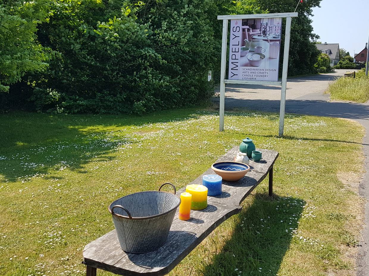 Holzbank am Wegesrand mit Kerzen und Keramik, dahinter ein Schild zur Werkstatt