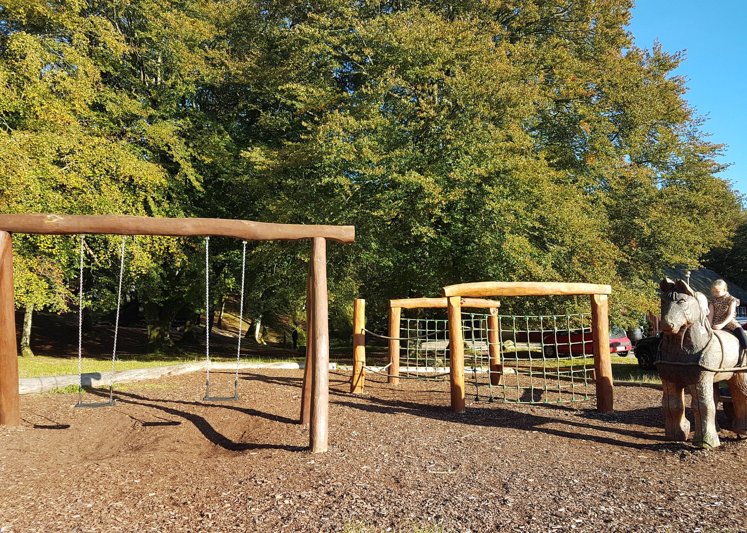 Spielplatz unter bunten Laubbäumen, rechts ein Kind auf einem Holzpferd