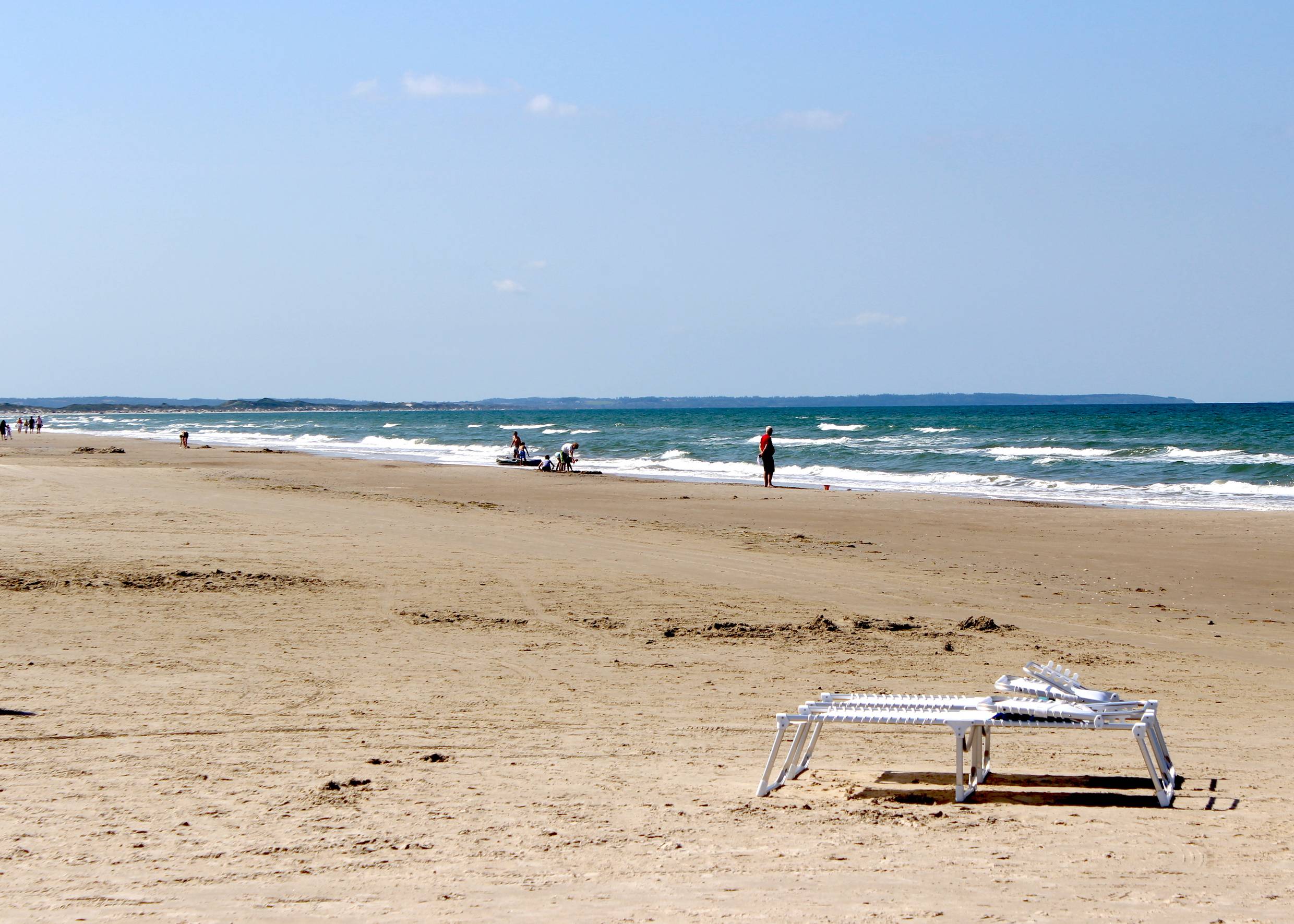 Breiter, langer Sandstrand am Meer, im Vordergrund eine Klappliege, am Wasser Badegäste
