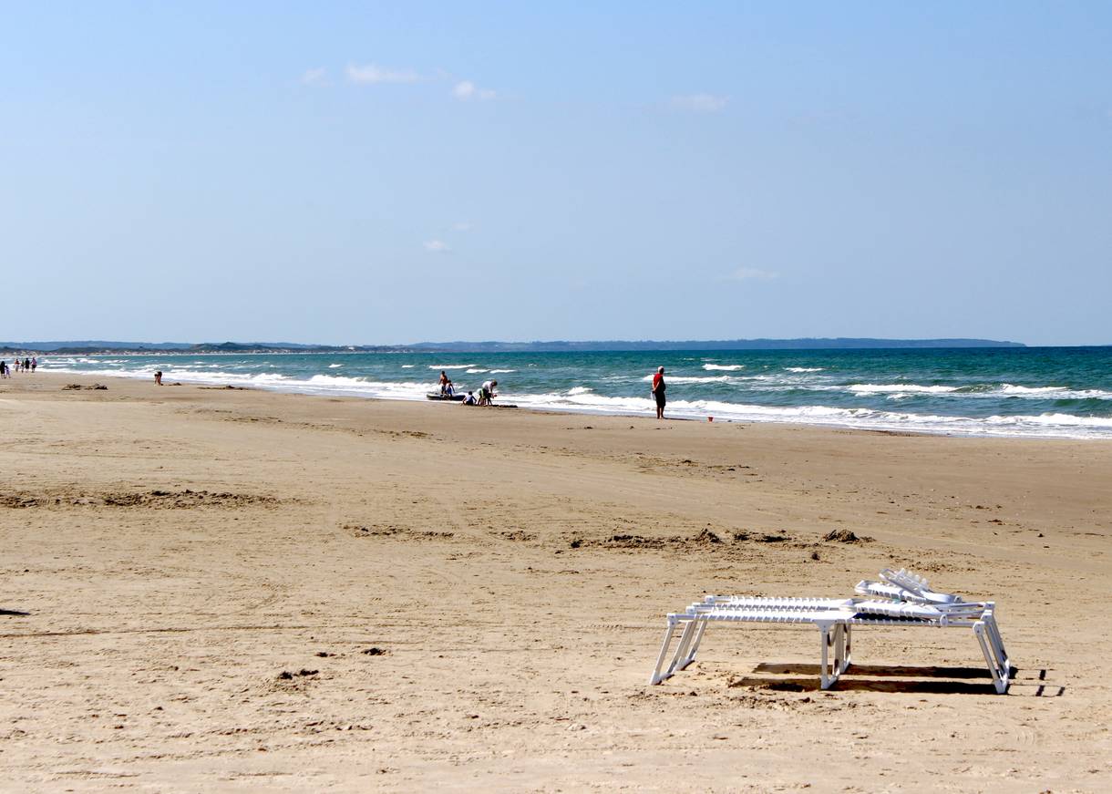 Breiter, langer Sandstrand am Meer, im Vordergrund eine Klappliege, am Wasser Badegäste