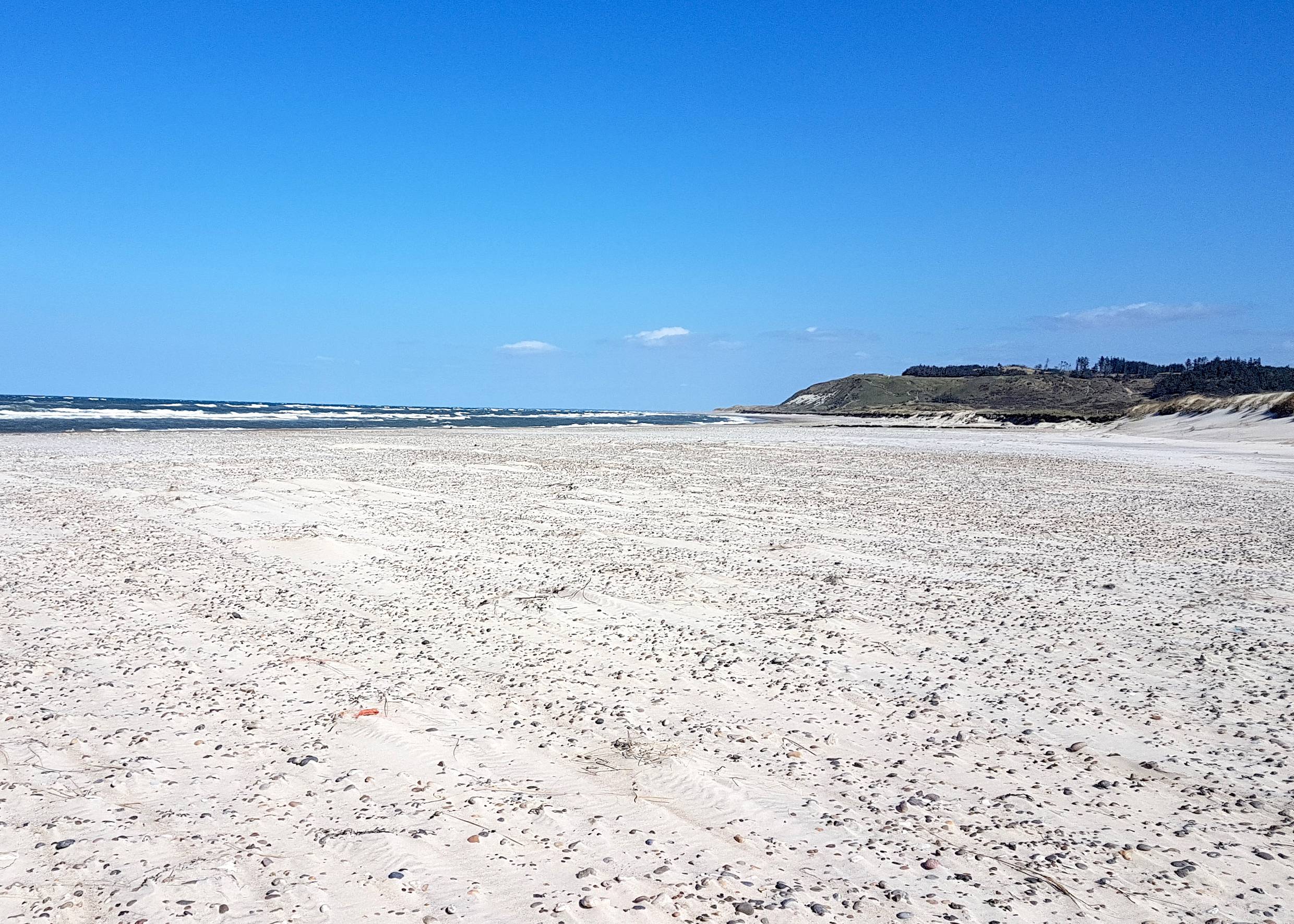 Steiniger Sandstrand unter blauem Himmel, im Hintergrund ein Hügel