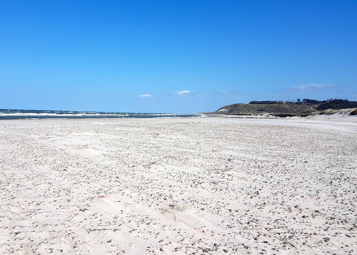Steiniger Sandstrand unter blauem Himmel, im Hintergrund ein Hügel