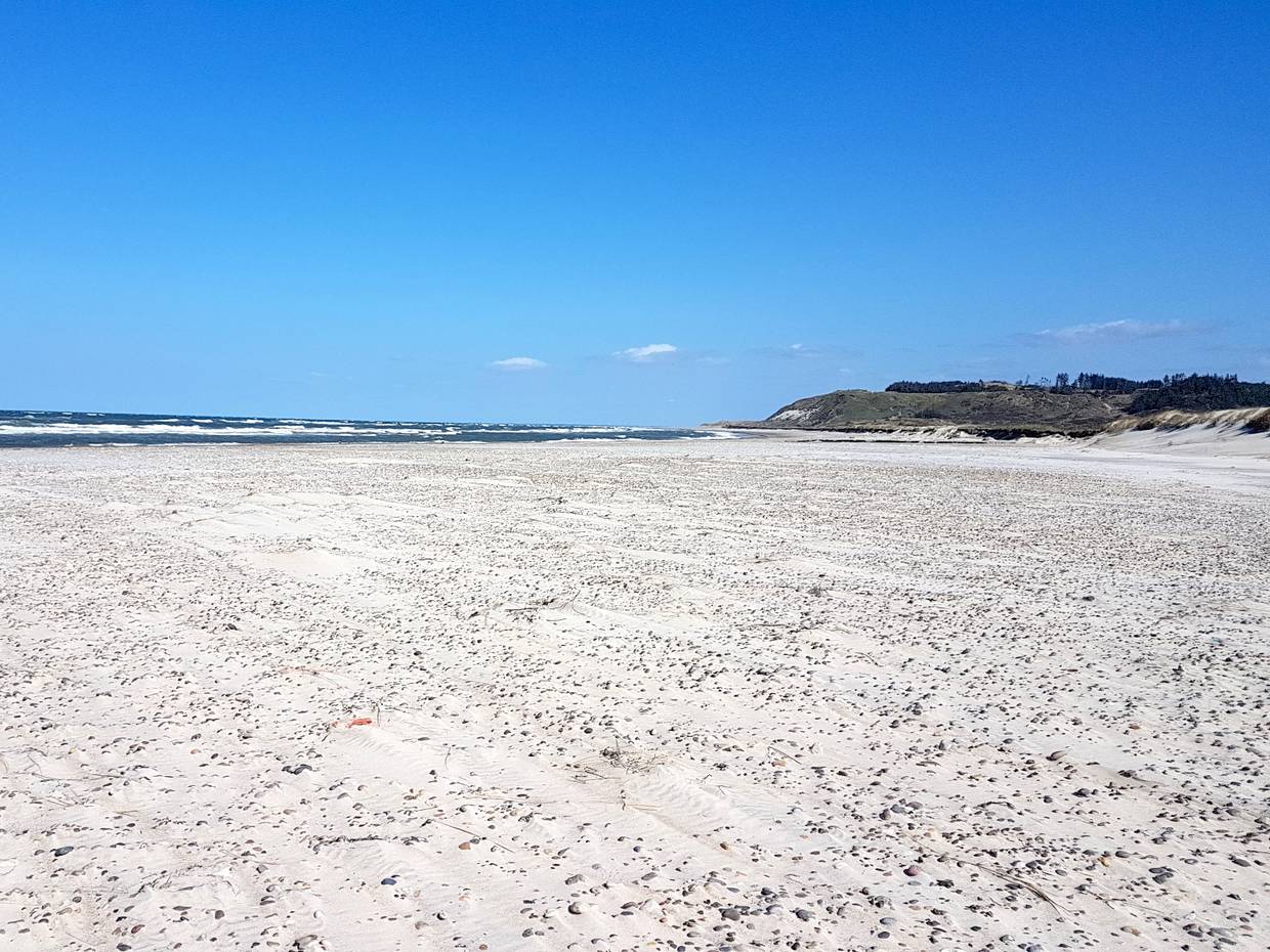 Steiniger Sandstrand unter blauem Himmel, im Hintergrund ein Hügel