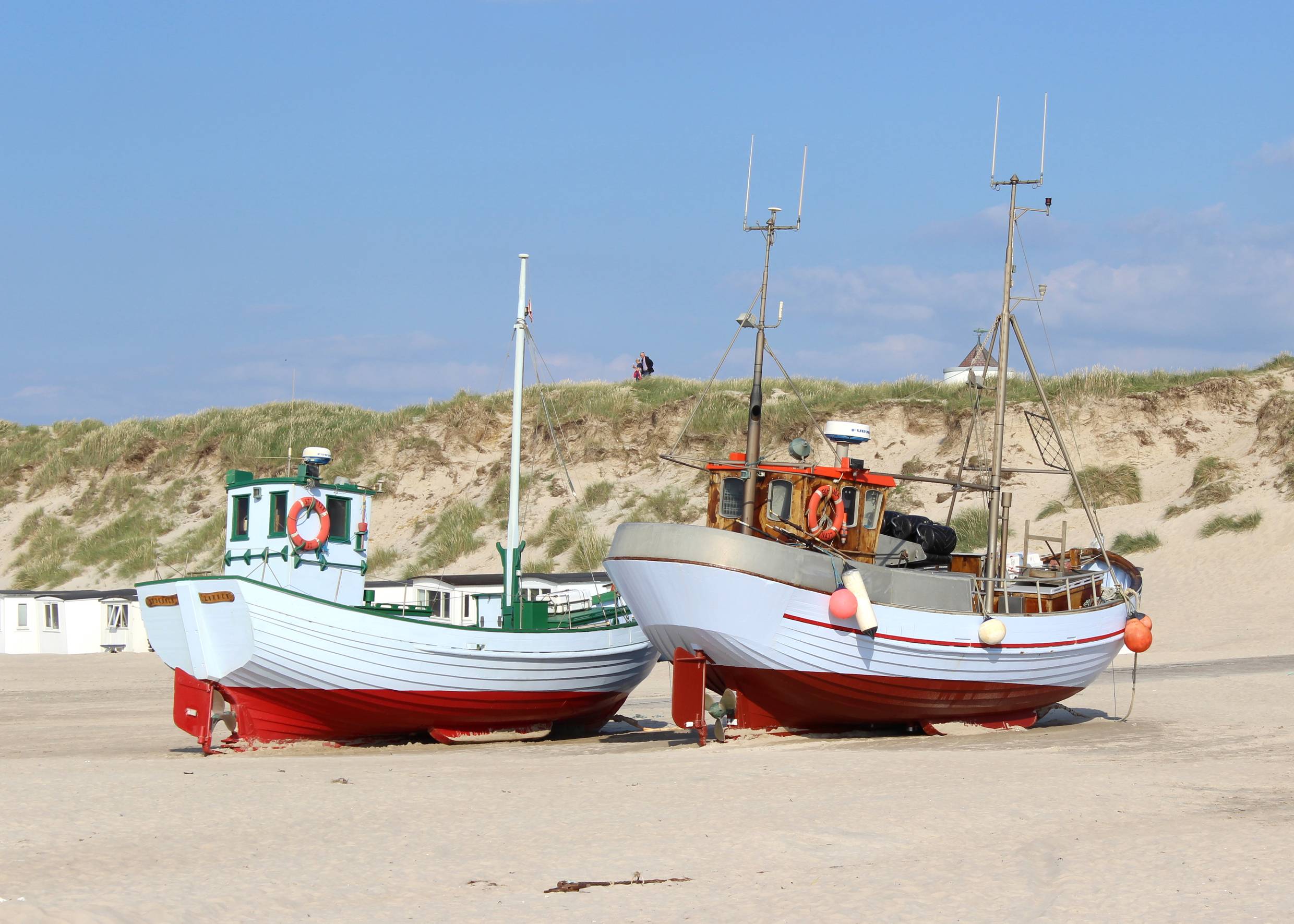 Hellblau-rotre Fischerboote auf dem Strand, dahinter Dünen und weiße Badehüttten